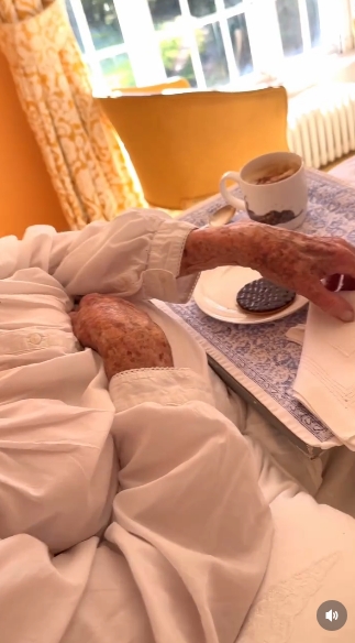 The camera shifts to a closer view of Lady Pamela Hicks's hands as she reaches forward, revealing delicate, time-worn features that speak to a life richly lived. The breakfast tray, complete with a neatly placed cup and saucer, adds to the quiet ritual of the moment. | Source: Instagram/indiahicksstyle