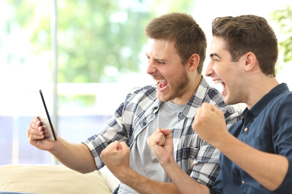 Dos emocionados amigos o compañeros de habitación viendo la televisión en línea en una tableta sentados en un sofá en la sala de estar en casa. I Foto: Shutterstock Dos emocionados amigos o compañeros de habitación viendo la televisión en línea en una tableta sentados en un sofá en la sala de estar en casa. I Foto: Shutterstock