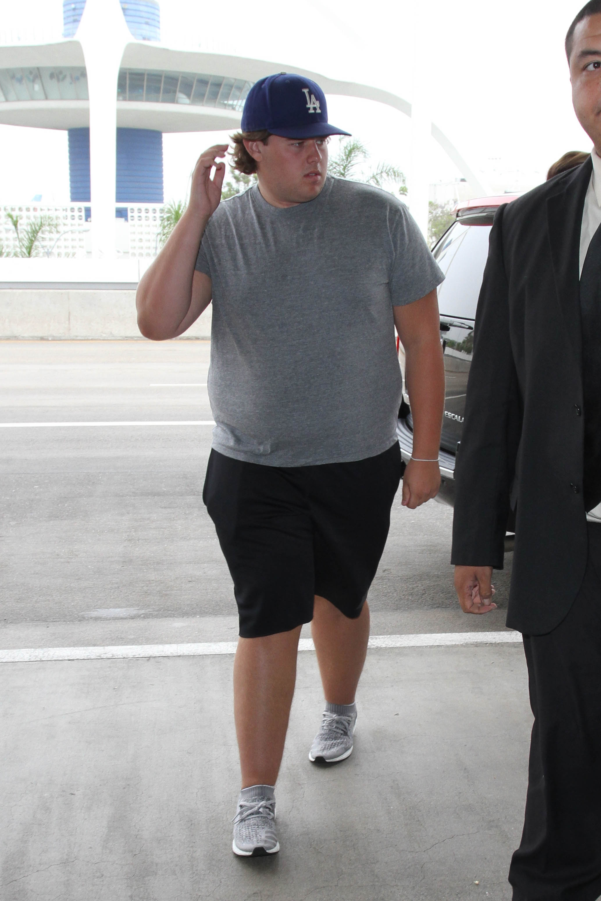 Christopher Schwarzenegger is seen at the airport in Los Angeles on August 31, 2016. Wearing a fitted grey t-shirt and a blue LA Dodgers cap, he carries a green backpack and military-style gear while navigating the airport with a heavier frame and visible focus on his belongings.