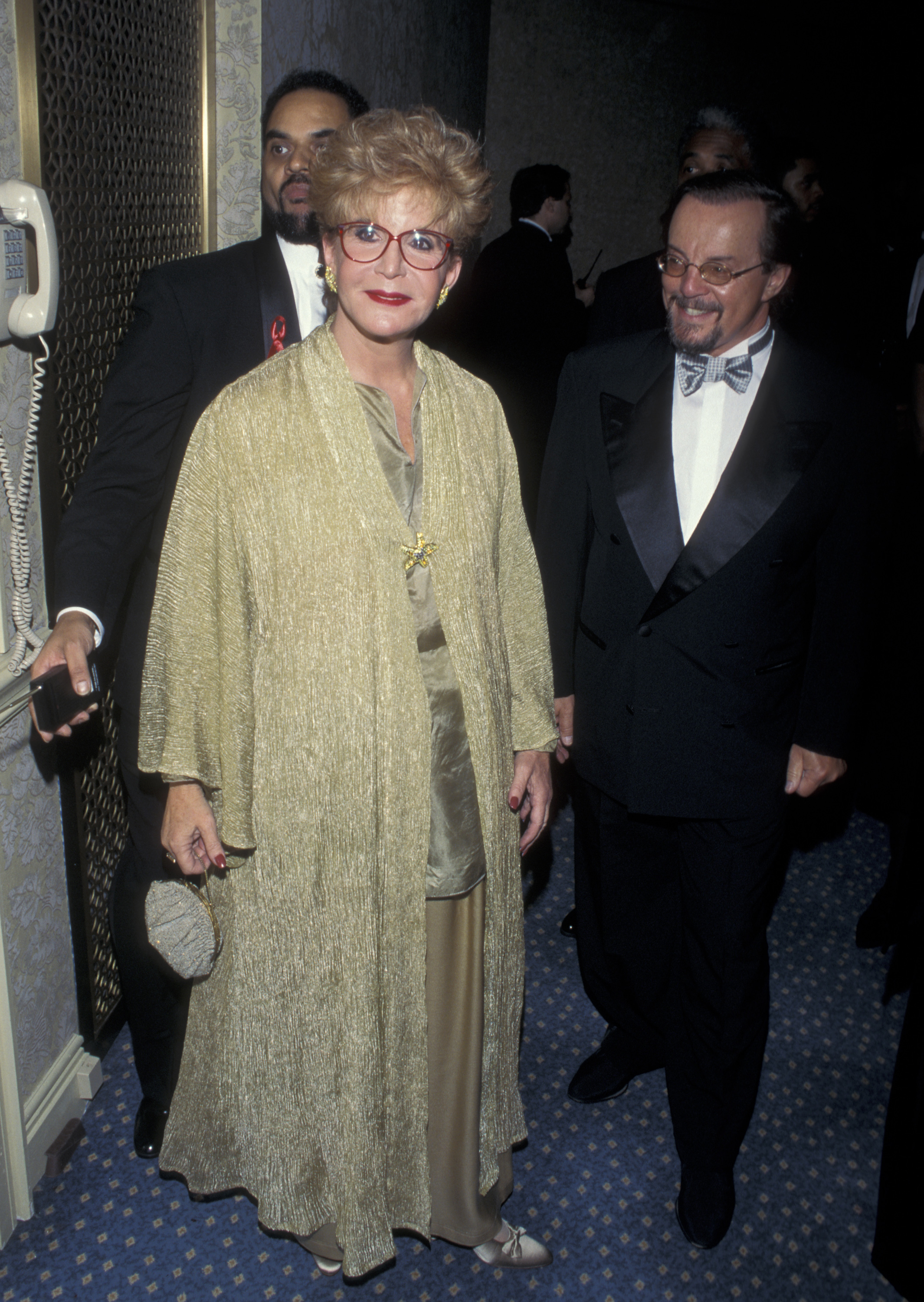 Sally Jessy Raphael and her husband, Karl Soderlund, attend the CORE Ambassadorial Reception Awards Dinner "Living the Dream" at the Sheraton Hotel in New York City on January 16, 1995. Raphael stands out in a shimmering gold ensemble and signature red frames, while Soderlund opts for a classic tuxedo with a bow tie.