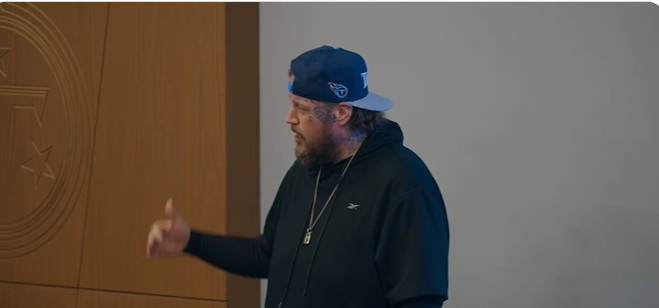 Jason "Jelly Roll" is seen mid-speech, wearing a navy blue Tennessee Titans cap backward and a black Reebok hoodie. A silver pendant necklace hangs around his neck as he gestures with his right hand, appearing engaged in conversation. His tattoos are partially visible, and the neutral indoor setting suggests a casual yet purposeful appearance. | Source:X/TennesseeTitans