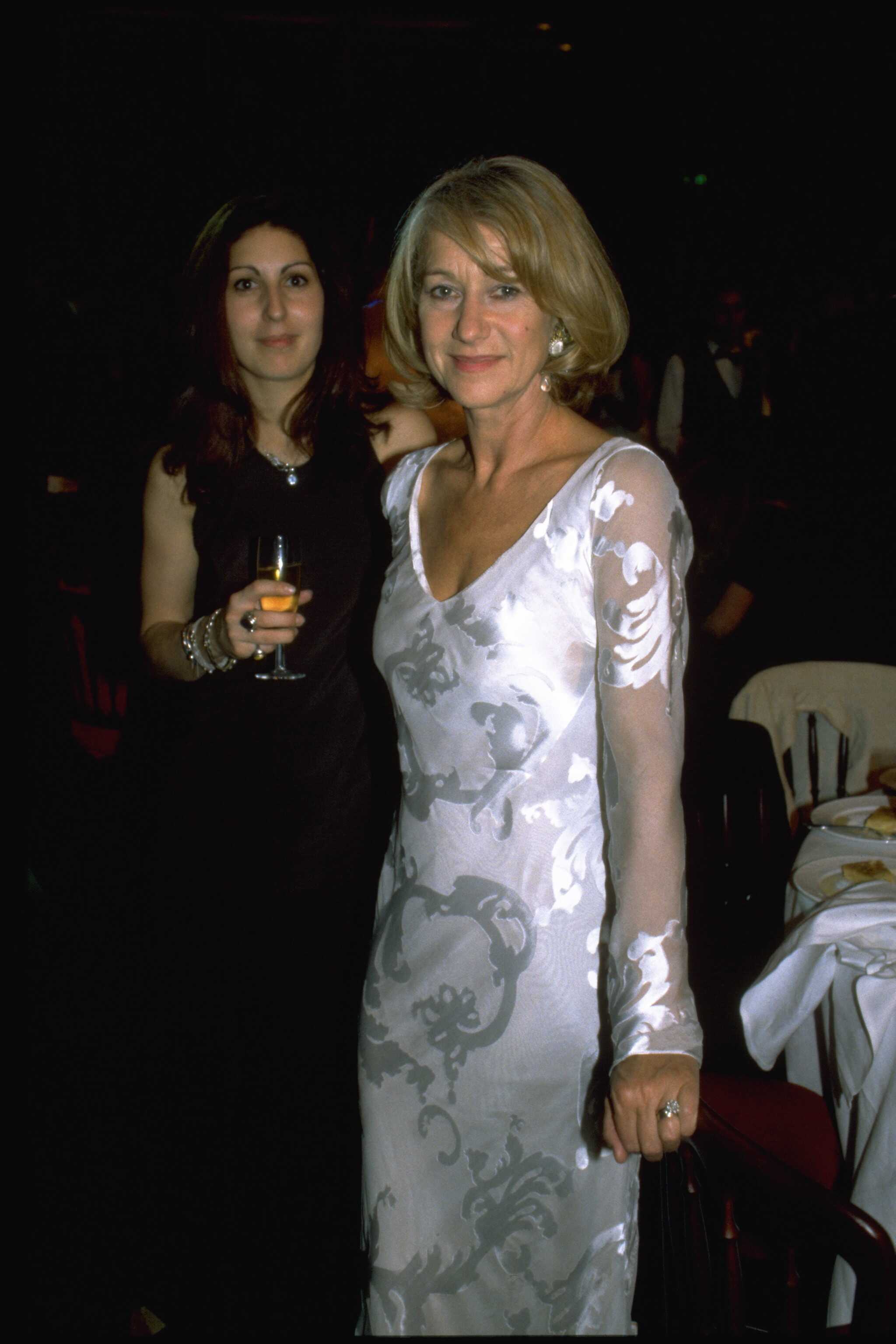 Helen Mirren dazzles at a formal evening event in a sheer, silver-patterned gown with a deep neckline and long sleeves, her look accented by elegant earrings and a soft blonde bob as she poses confidently beside a guest holding a champagne flute.