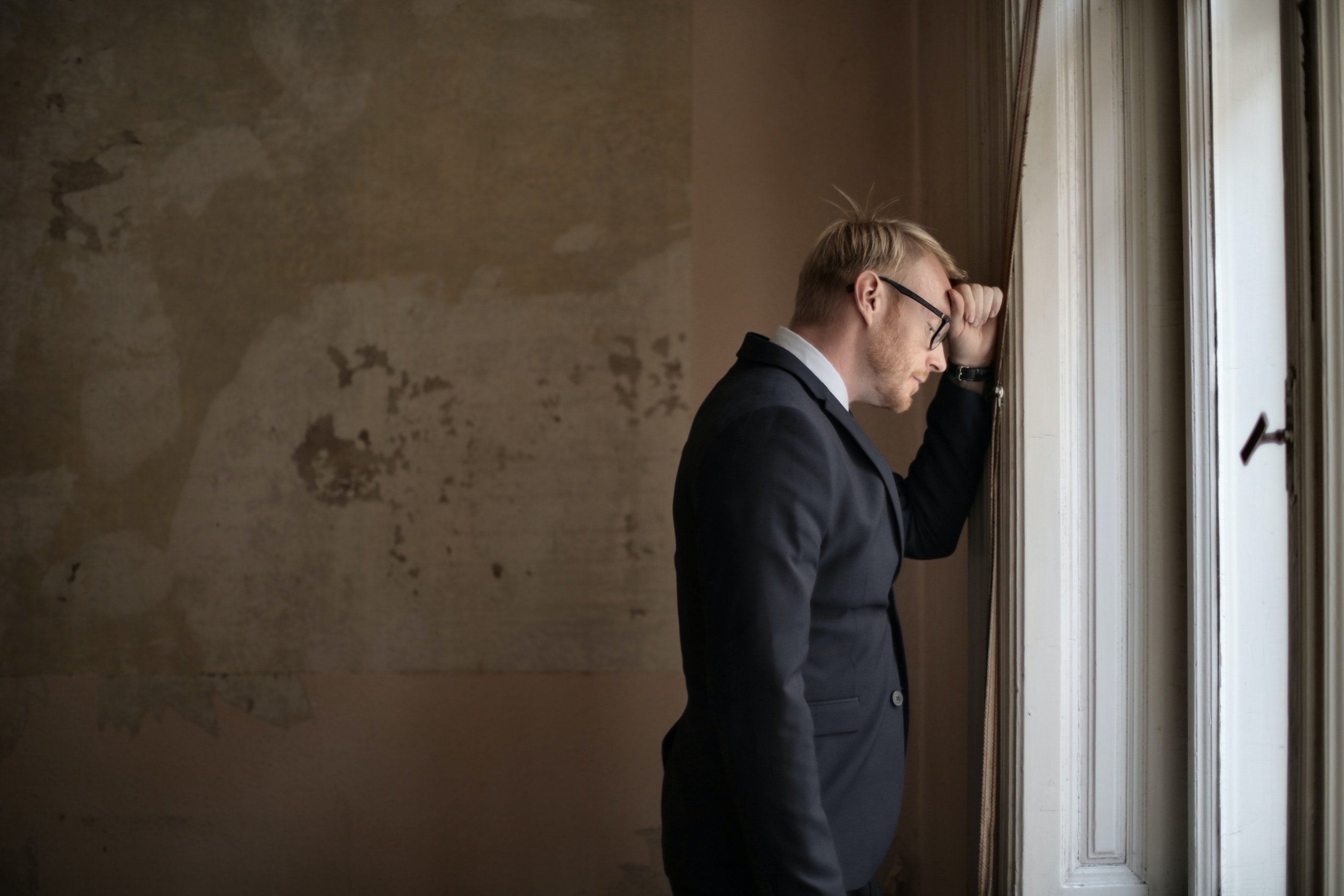 Man in a suit looking out a window with his head down and his hand on his forehead. | Photo: Pexels