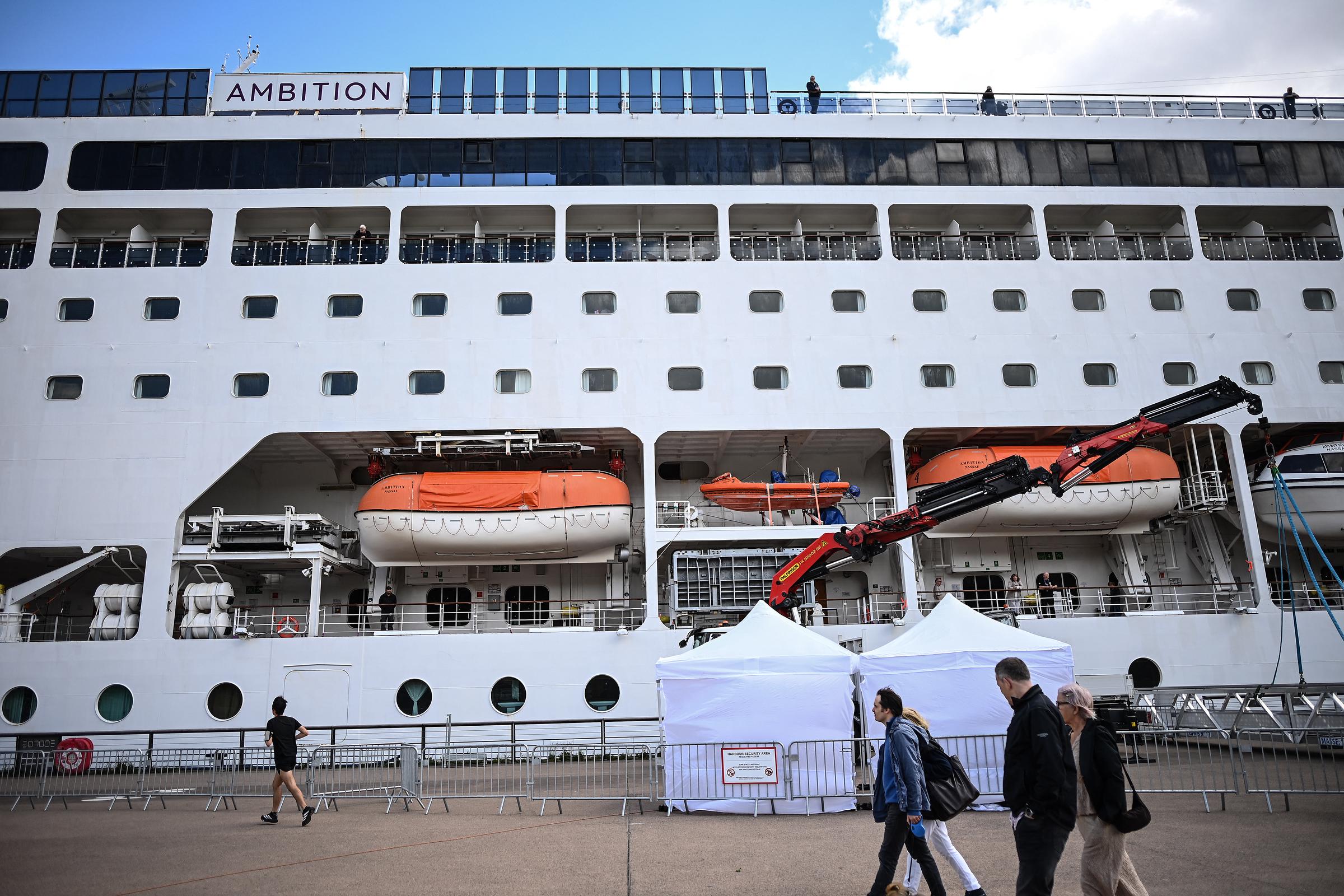 Passers-by walk past the Bahamas-registered cruise ship Ambition after passengers were confined following the outbreak of a gastrointestinal illness onboard, at the Bordeaux port in Bordeaux, southwestern France on May 13, 2026. | Source: Getty Images