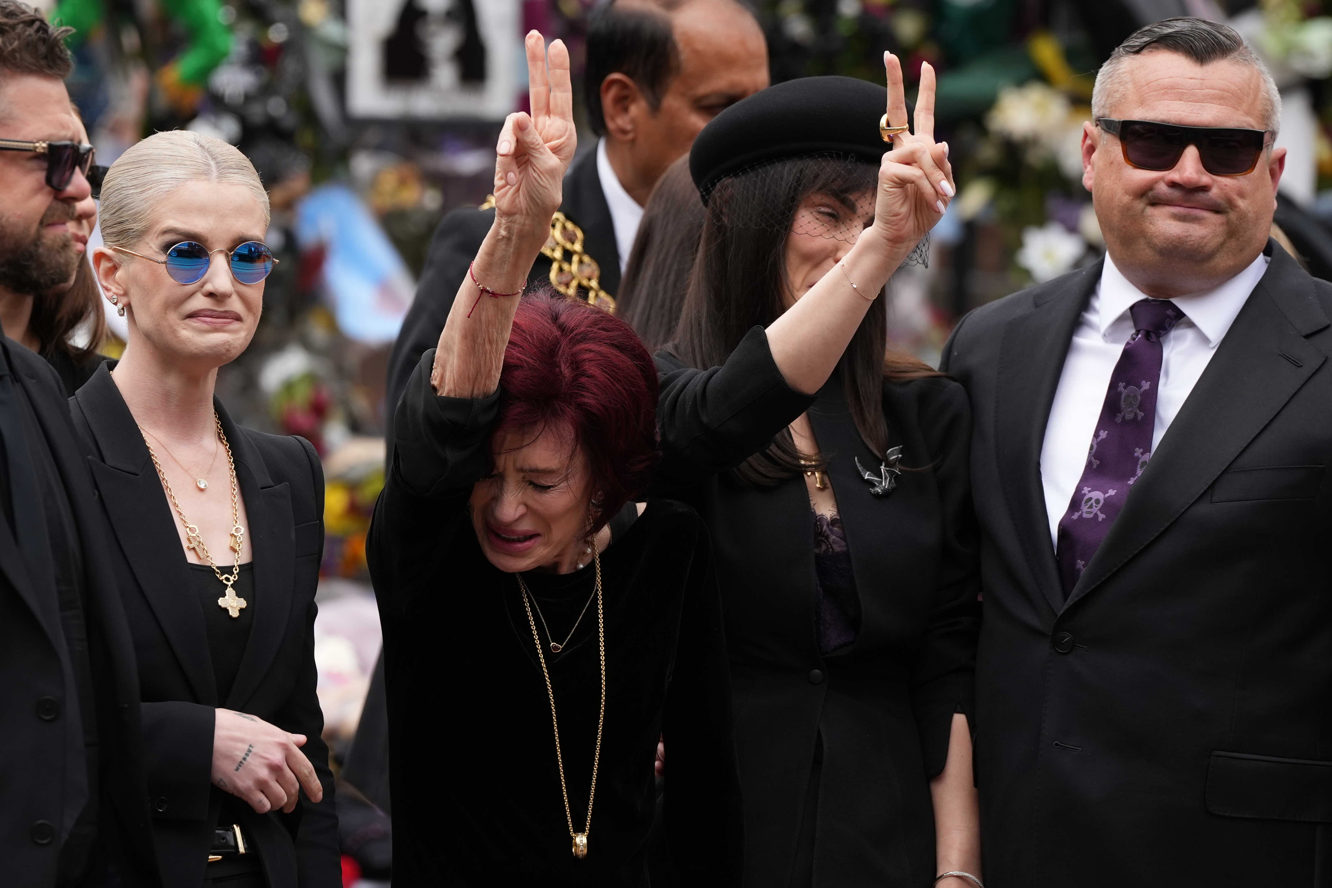 Sharon Osbourne mourns during the funeral ceremony of her late husband, Ozzy. Their children Jack, Kelly, and Aimee also mourn their father. The whole family is overcome with grief. Aimee and Sharon hold up the peace sign while Jack and Kelly are seen crying.