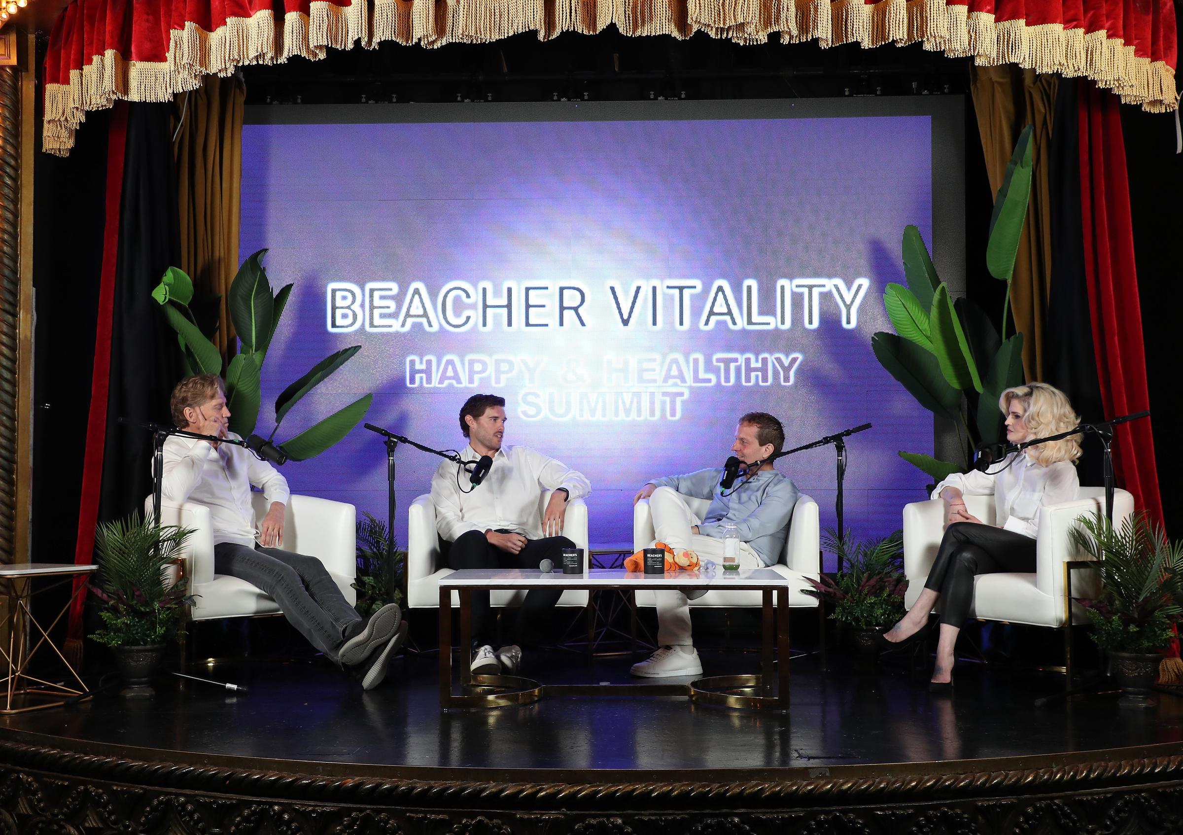 Christopher Schwarzenegger is seen at the Beacher Vitality Happy & Healthy Summit on May 10, 2025. Seated beside Dr. Robert Huizenga, Jeff Beacher, and Kelly Osbourne, he engages in a conversation focused on health and transformation, dressed in a crisp white shirt and black pants.