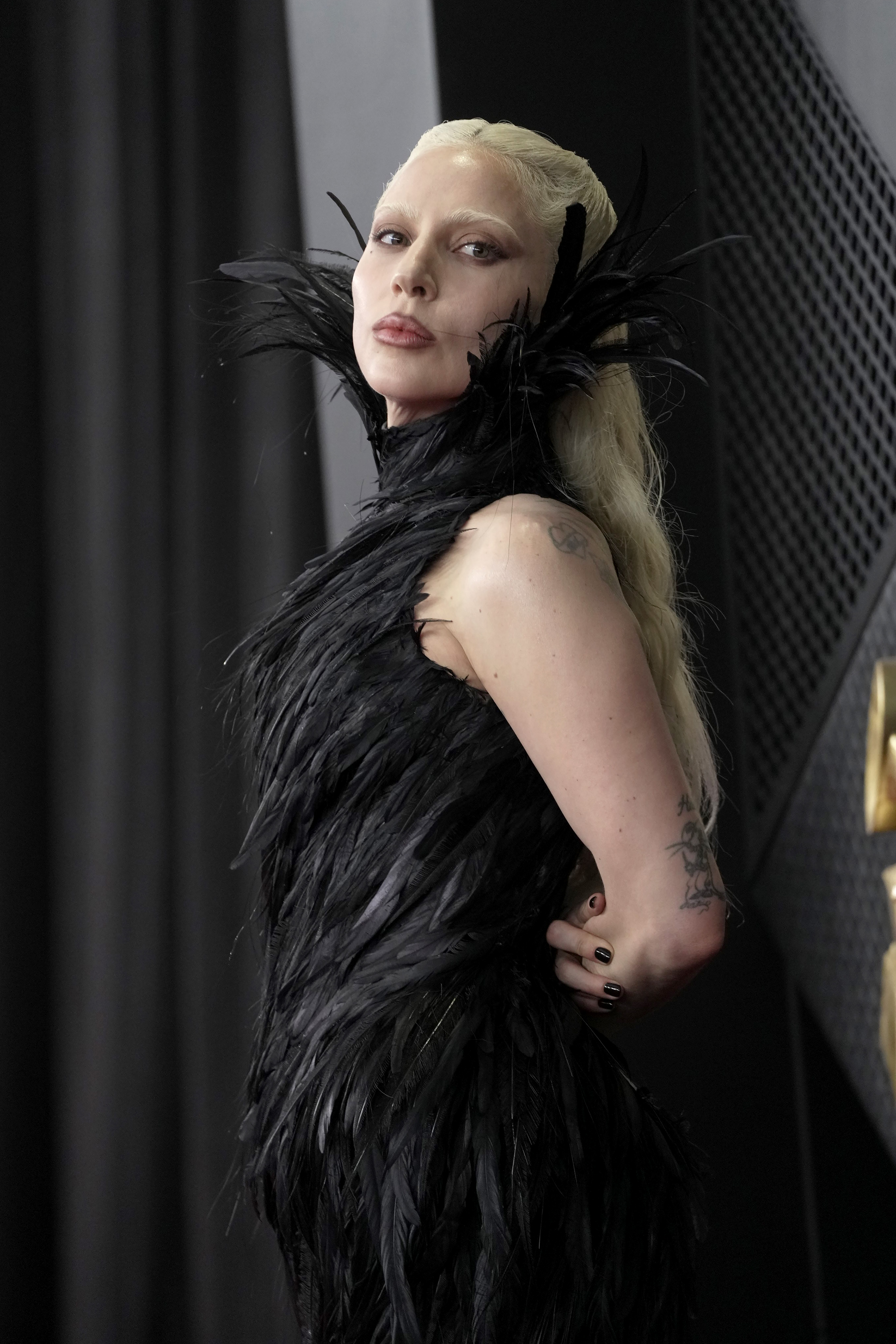 Lady Gaga poses on the red carpet at the 68th Grammy Awards at Crypto.com Arena in Los Angeles, California, wearing a black, feathered ensemble that wrapped around her shoulders and upper body. The look featured dense, layered plumage that created a dramatic silhouette, contrasting with exposed skin along her arms and neckline.
