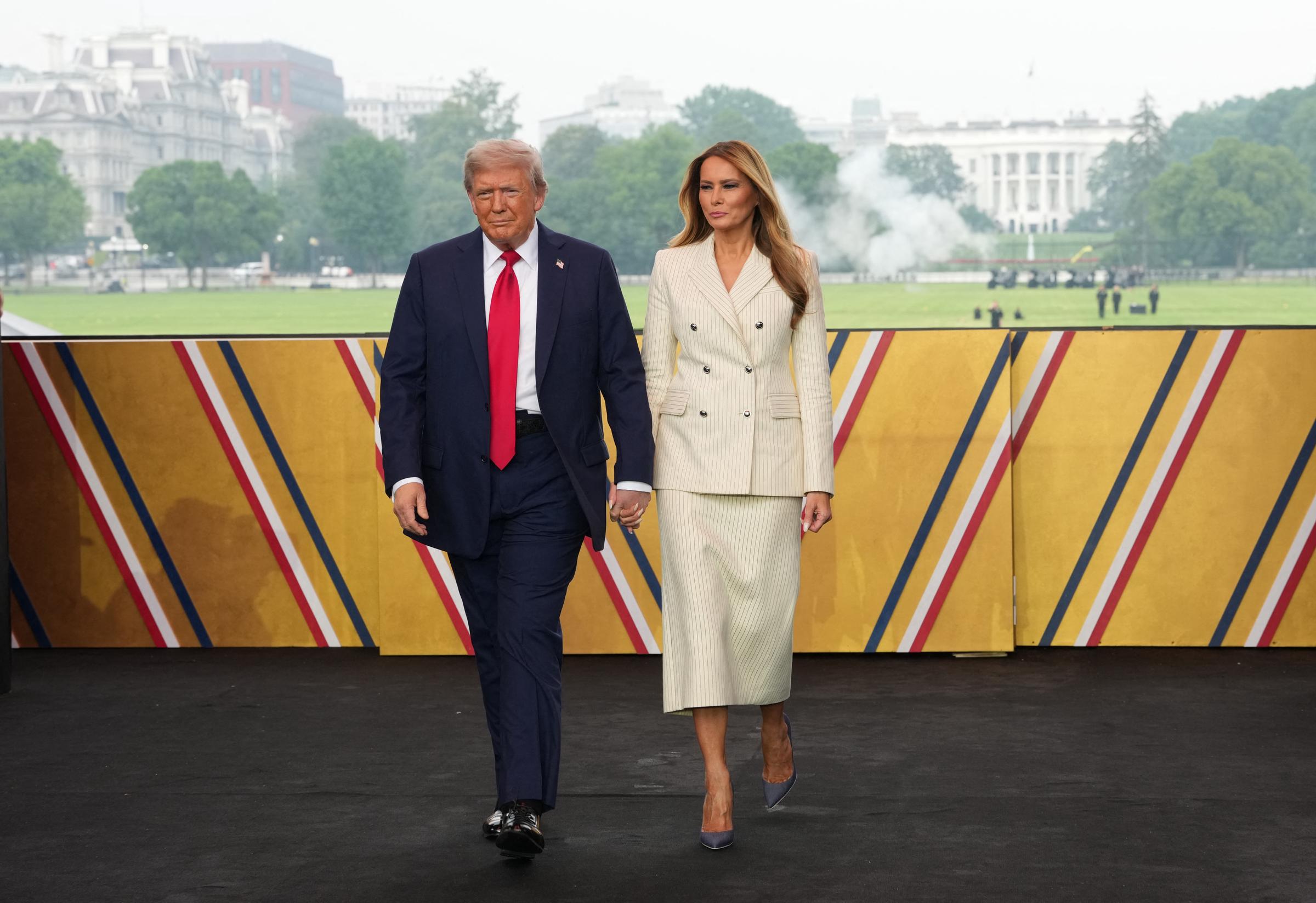 Donald and Melania Trump walk hand in hand toward the stage, with the White House and parade festivities in the background. Melania pairs her top with a cream pinstripe skirt suit as they make their entrance against a patriotic backdrop.