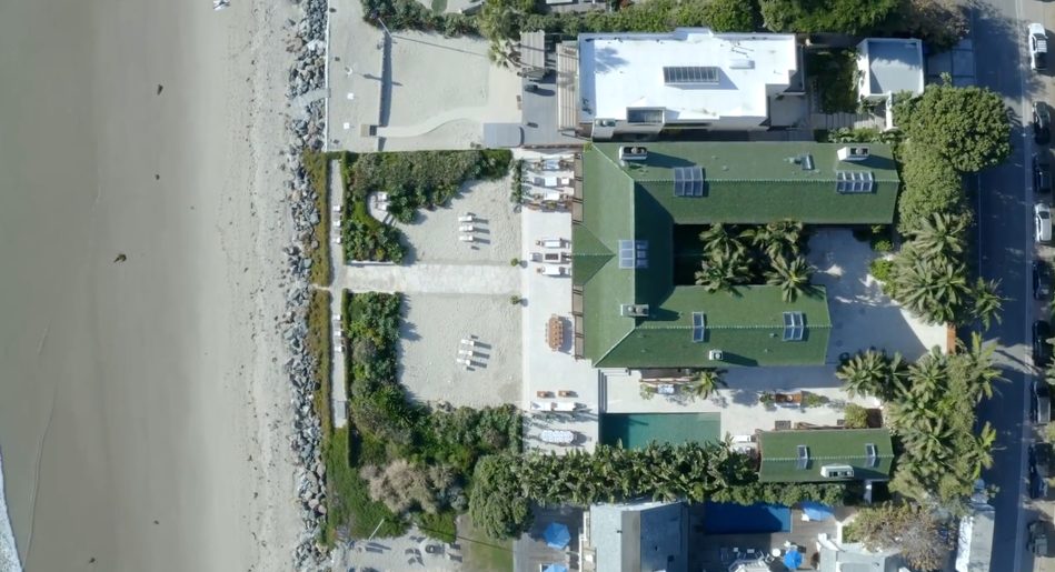 An overhead aerial shot reveals the full footprint of Orchid House, highlighting its U-shaped green roof structure centered around an inner tropical courtyard. The property stretches directly to the shoreline, complete with a private sandy lounge area, adjacent pool, and immediate access to the beach. | Source: YouTube/Architectural Digest