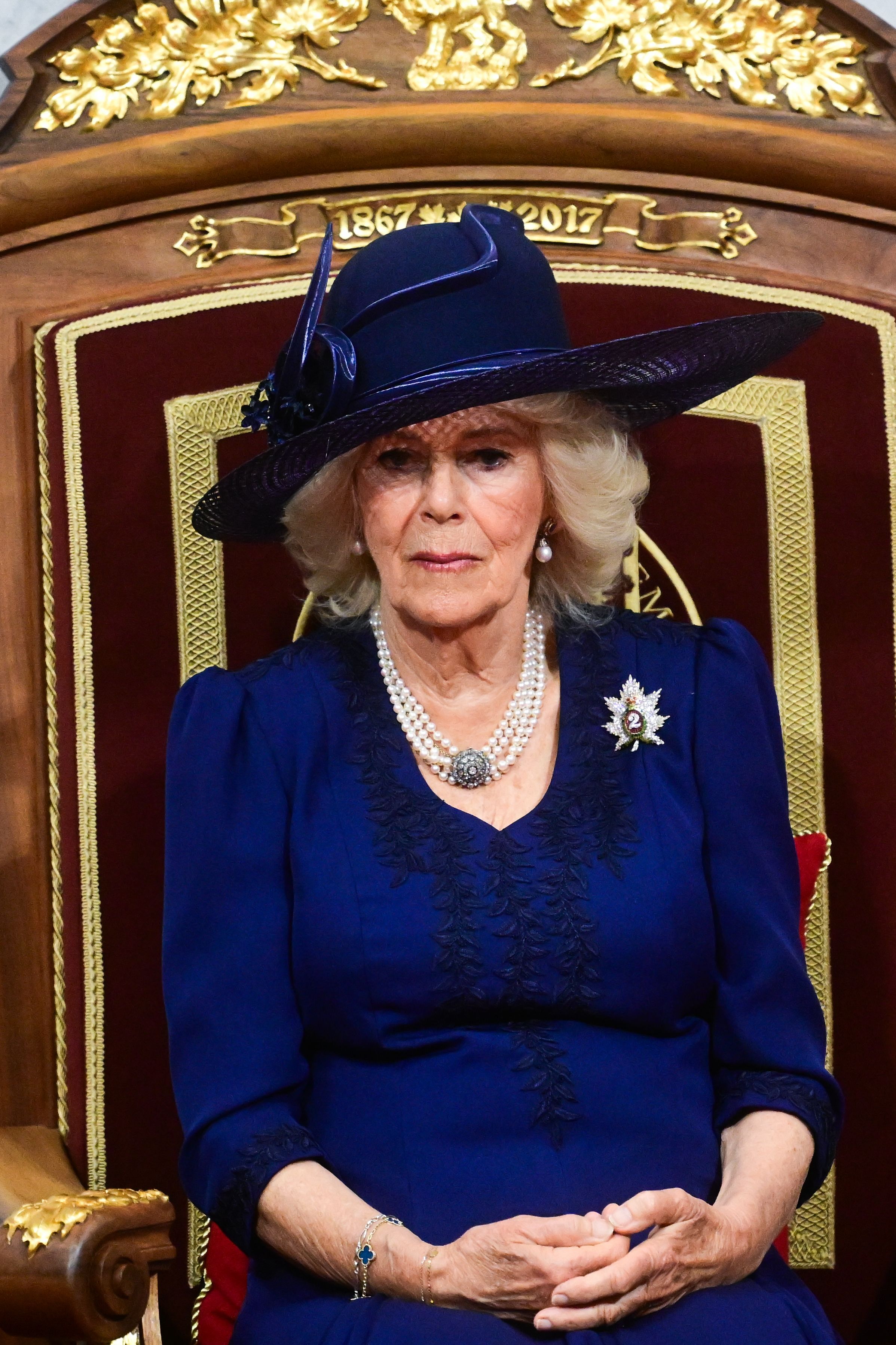 Queen Camilla, seated, wears a navy blue dress with a matching hat for the second day of the Canadian visit. She accessorizes with her signature four-row pearl choker, diamond and pearl drop earrings, and a badge from the Queen's Own Rifles of Canada.