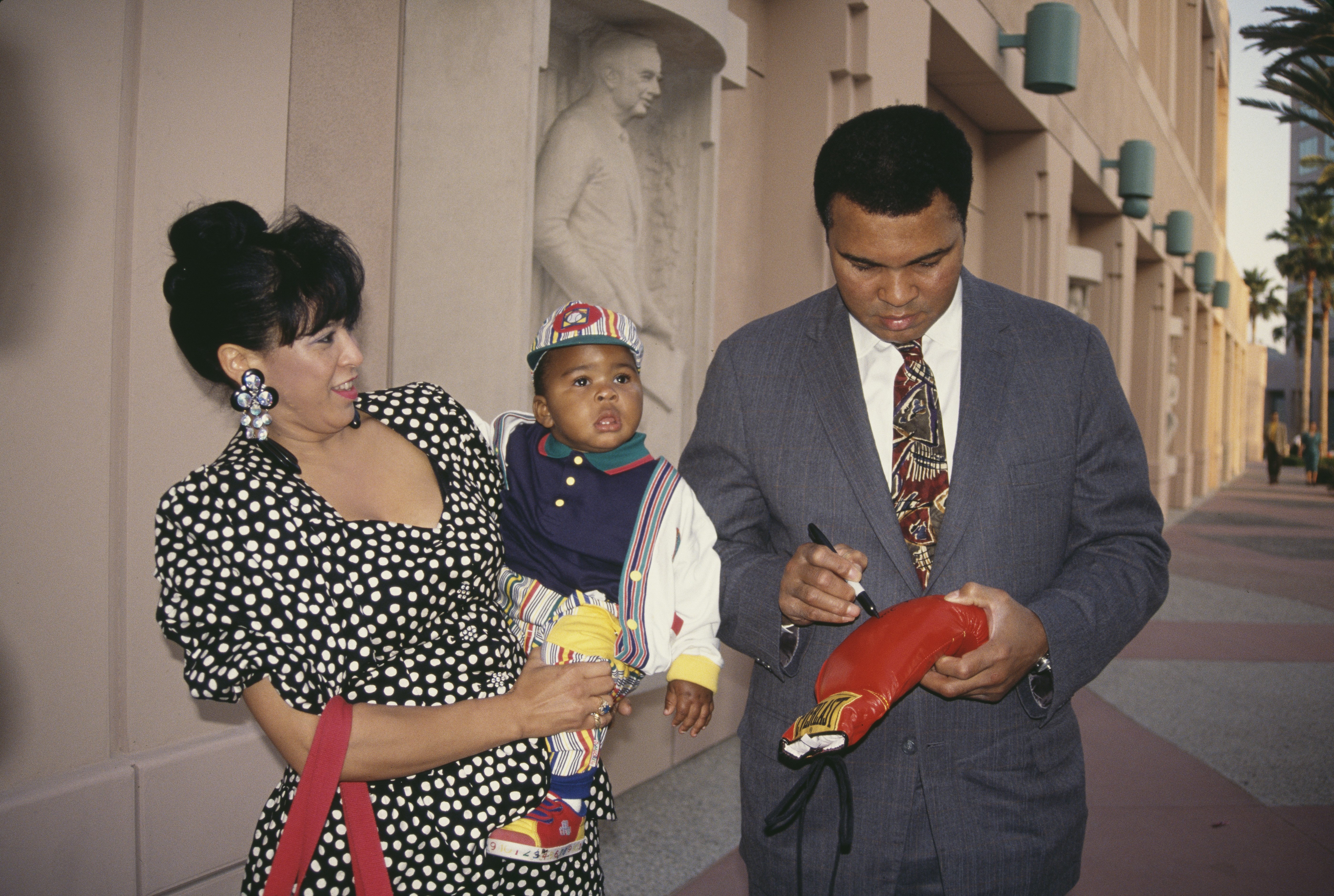 Yolanda "Lonnie" Ali holding her adopted son, Asaad Ali, as husband, American heavyweight boxer Muhammad Ali autographs a boxing glove, circa 1986. | Source: Getty Images