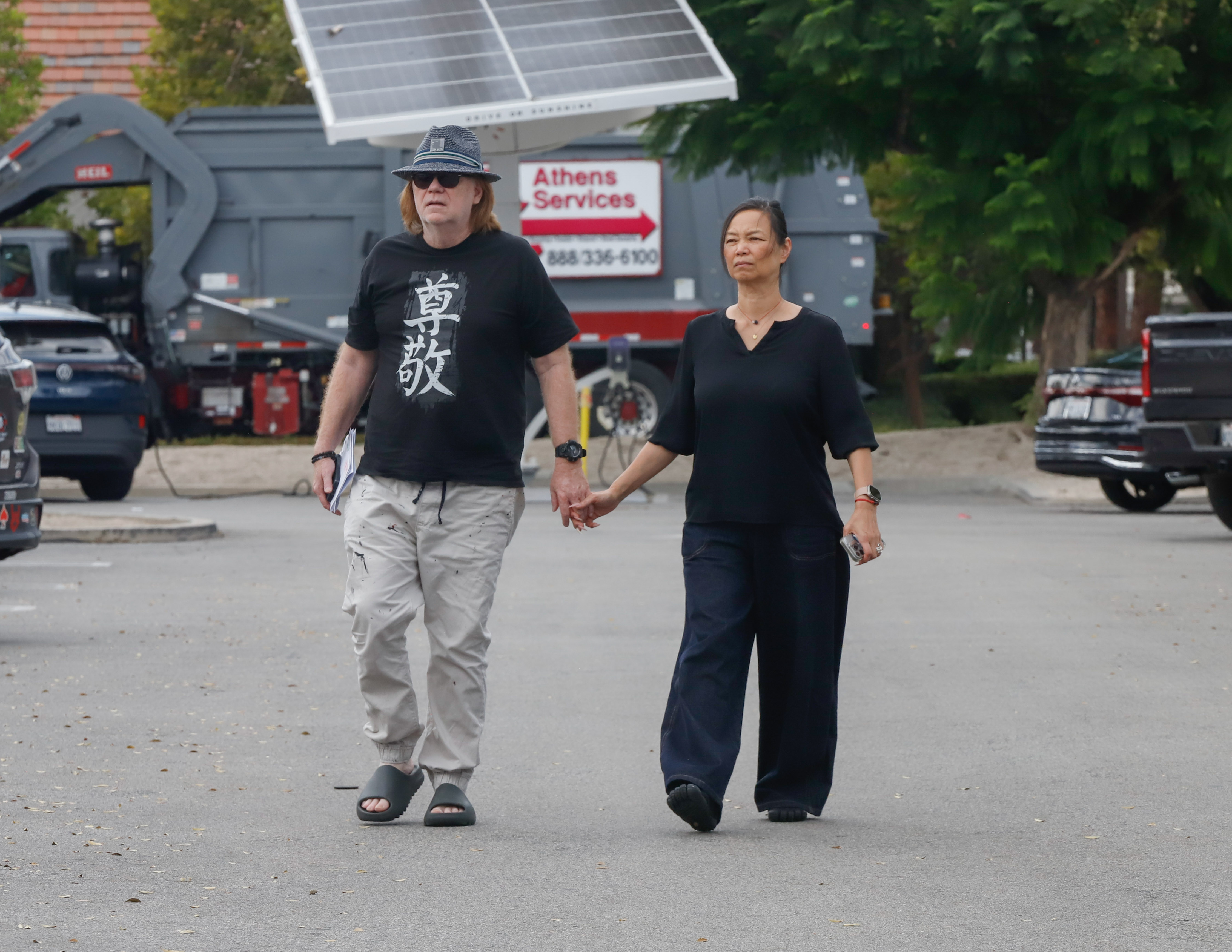 David Caruso walks hand in hand with a woman while running errands in Los Angeles, wearing sunglasses, a hat, a black T-shirt, light pants, and sandals.