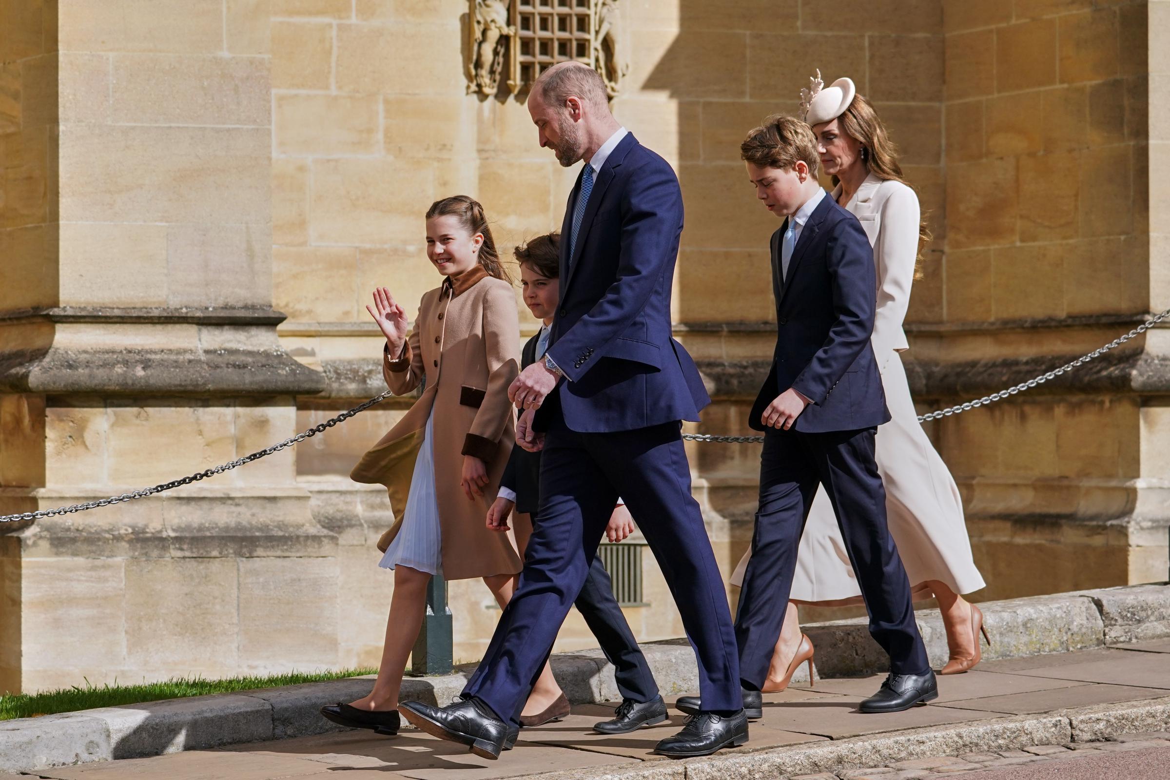 Shot in profile along the sun-drenched exterior of St George's Chapel, this image shows the Wales family in full stride, the Prince of Wales leading the group with Prince Louis tucked close beside him. Princess Charlotte waves cheerfully to onlookers on the left, her blue pleated dress lifting in the breeze beneath her camel coat, while Prince George and the Princess of Wales follow just behind.