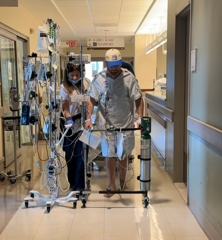 Daniel Neeson walks through a hospital hallway in a gown and cap, supported by medical equipments and a staff member beside him. | Source: Instagram/bgdans91