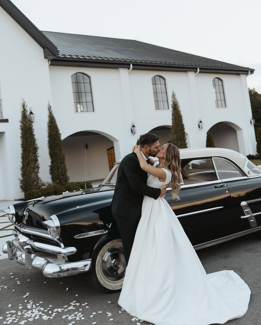 Jana Duggar and Stephen Wissmann share a kiss beside a vintage black car, her flowing gown sweeping the ground as flower petals scatter around them. | Source: Instagram/janamduggar