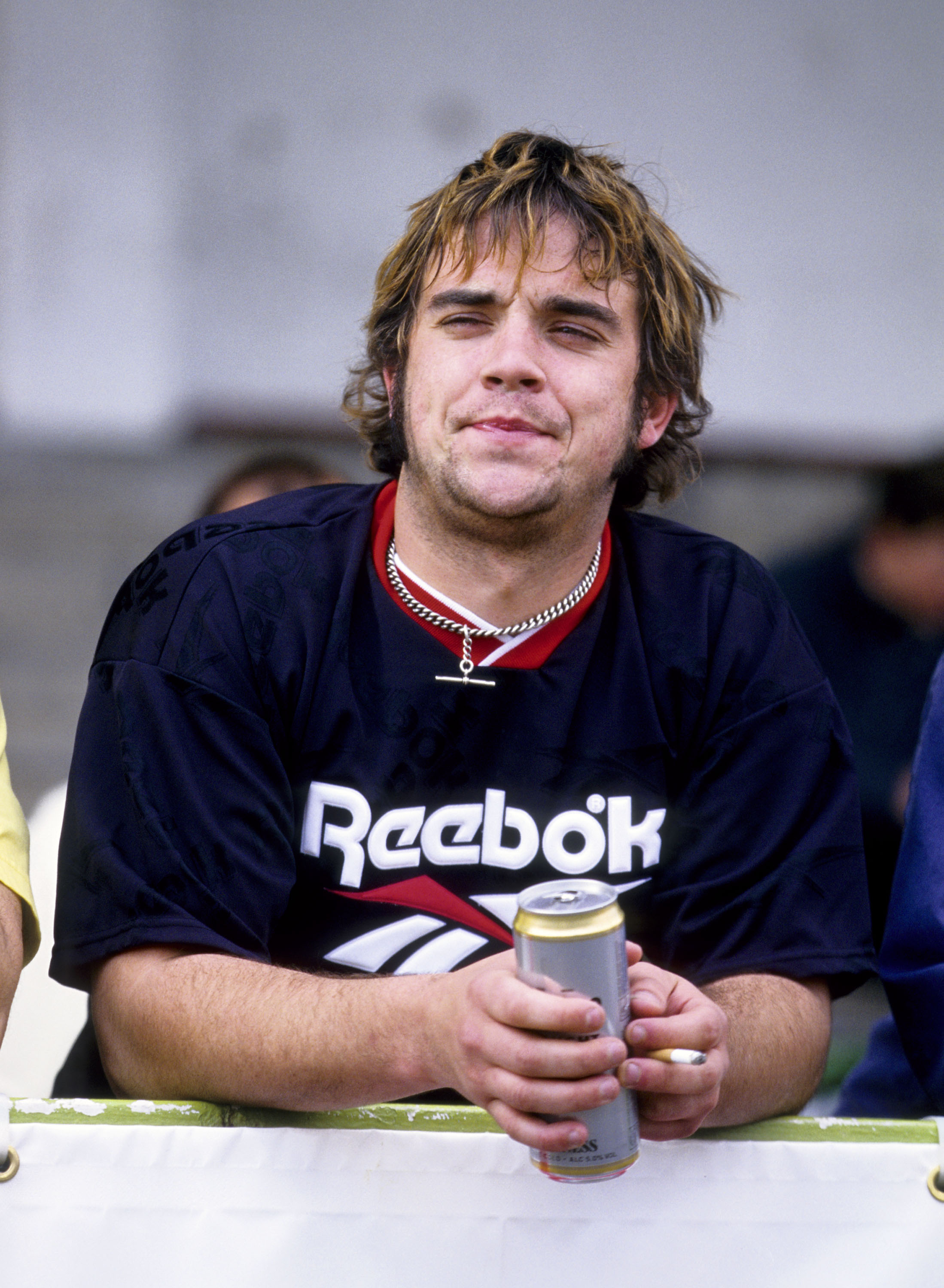 Robbie Williams attended a charity soccer match in May 1996, holding a beer and a cigarette while wearing a Reebok jersey. The candid moment reflected the rebellious image he embraced after leaving Take That.