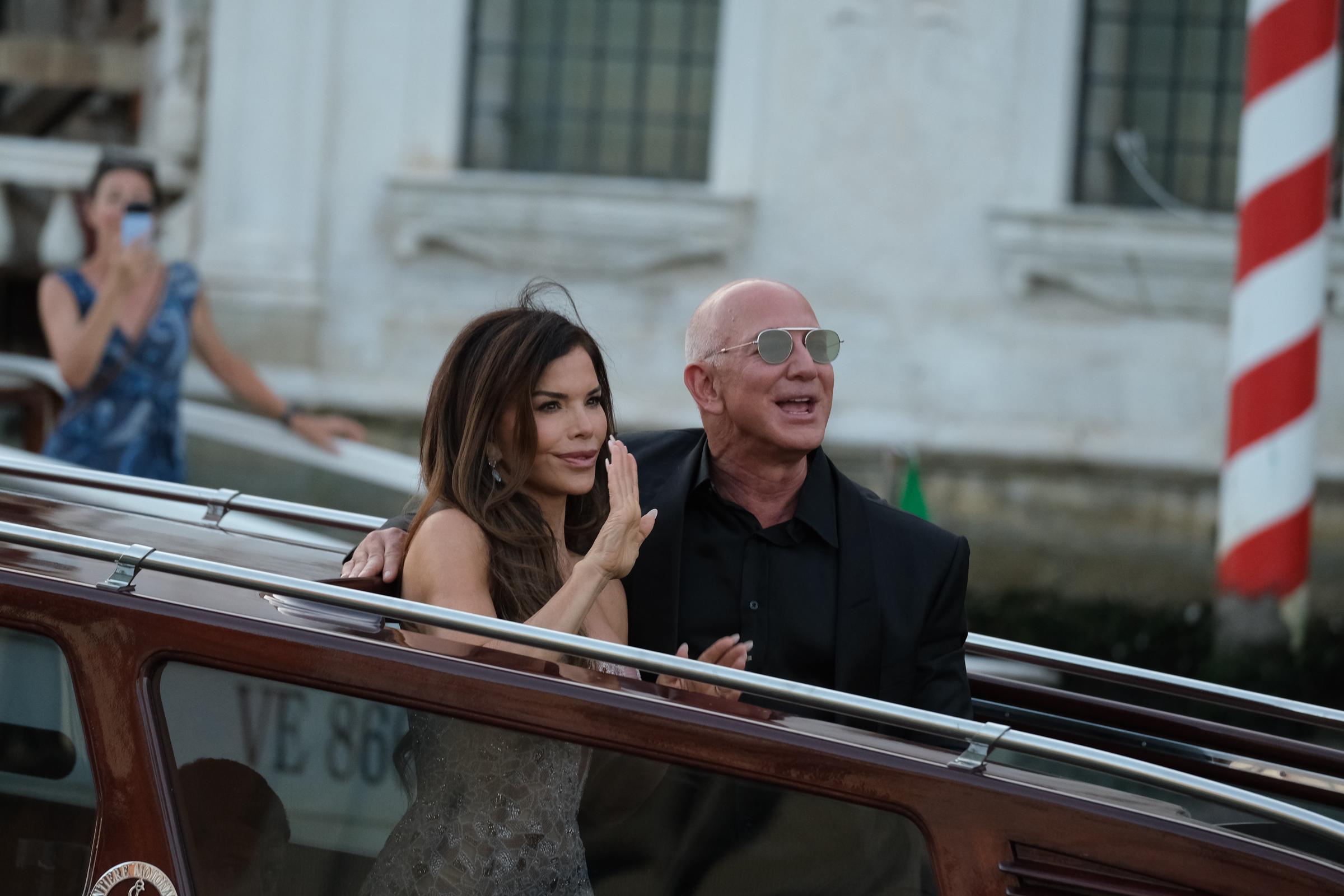 Lauren Sánchez raises her hand in a gentle wave as Jeff Bezos smiles beside her, both standing on the boat while a woman in the background captures the moment on her phone near a red-and-white striped post.