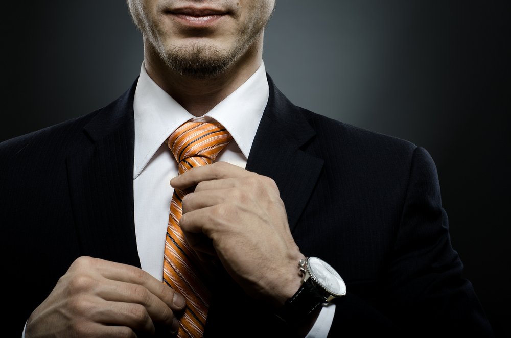 A portrait of a man dressed in a black suit while putting on an orange striped necktie | Photo: Shutterstock/Aleksey Mnogosmyslov