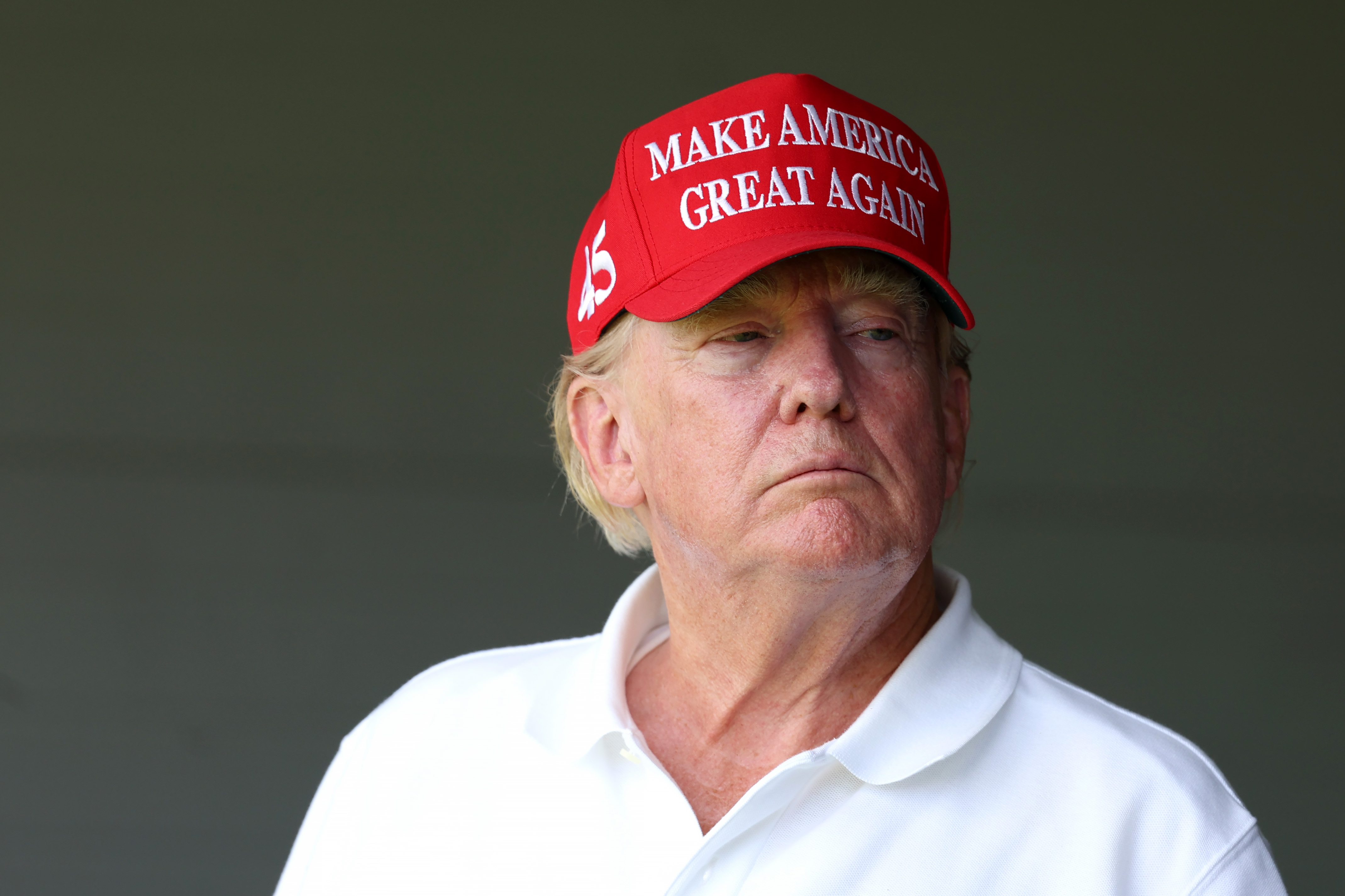 On May 26, Donald Trump watches from a shaded area wearing the same red hat and white shirt. His skin tone looks more even in the dimmer light, though the red coloring is still strong near his cheeks.