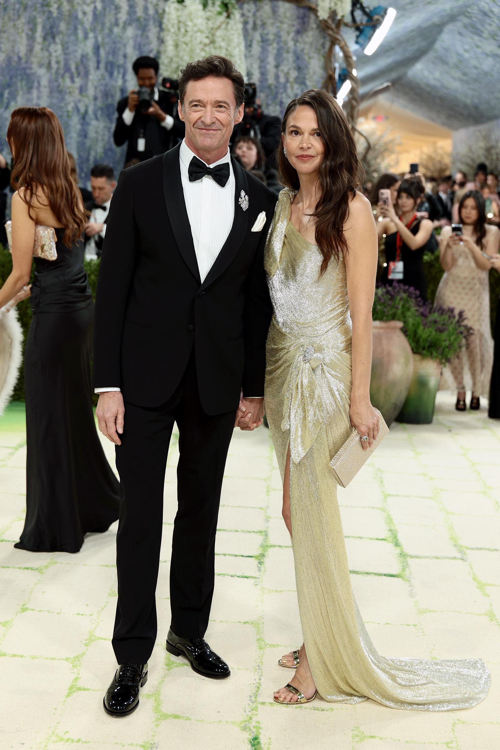 Hugh Jackman offers a polished smile in a classic black tuxedo, crisp bow tie, and glossy shoes that reflect the flash of cameras. Hand in hand, Sutton Foster glows in a draped gold gown with a subtle shimmer, her soft waves and minimal jewelry letting the fluid silhouette do all the talking.