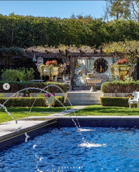 The rear garden frames the mosaic-tiled pool in full, with arching water jets catching the California sun and clipped box hedging lining a stone pathway toward a pergola. Oversized antique urns spill with flowers on either side of a large ornamental clock mounted against the back wall, the whole scene carrying the quietly theatrical sensibility that ran through every corner of the Osbourne estate. | Source: Instagram/top.music.songs