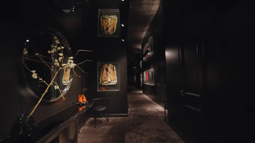 A dramatic, dimly lit hallway in the basement featuring matte black walls, a large circular mirror, and museum-style acrylic cases displaying iconic stage outfits. | Source: YouTube/@Archdigest