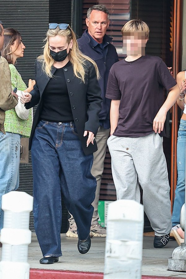 Adele, dressed in wide-leg dark denim, a black jacket, and ballet flats, walks closely alongside her teenage son Angelo — who sports grey sweatpants and a dark brown tee — as they navigate their way through a crowd outside South Beverly Grill, with a suited security guard following closely behind. | Source: X/Pop Tingz