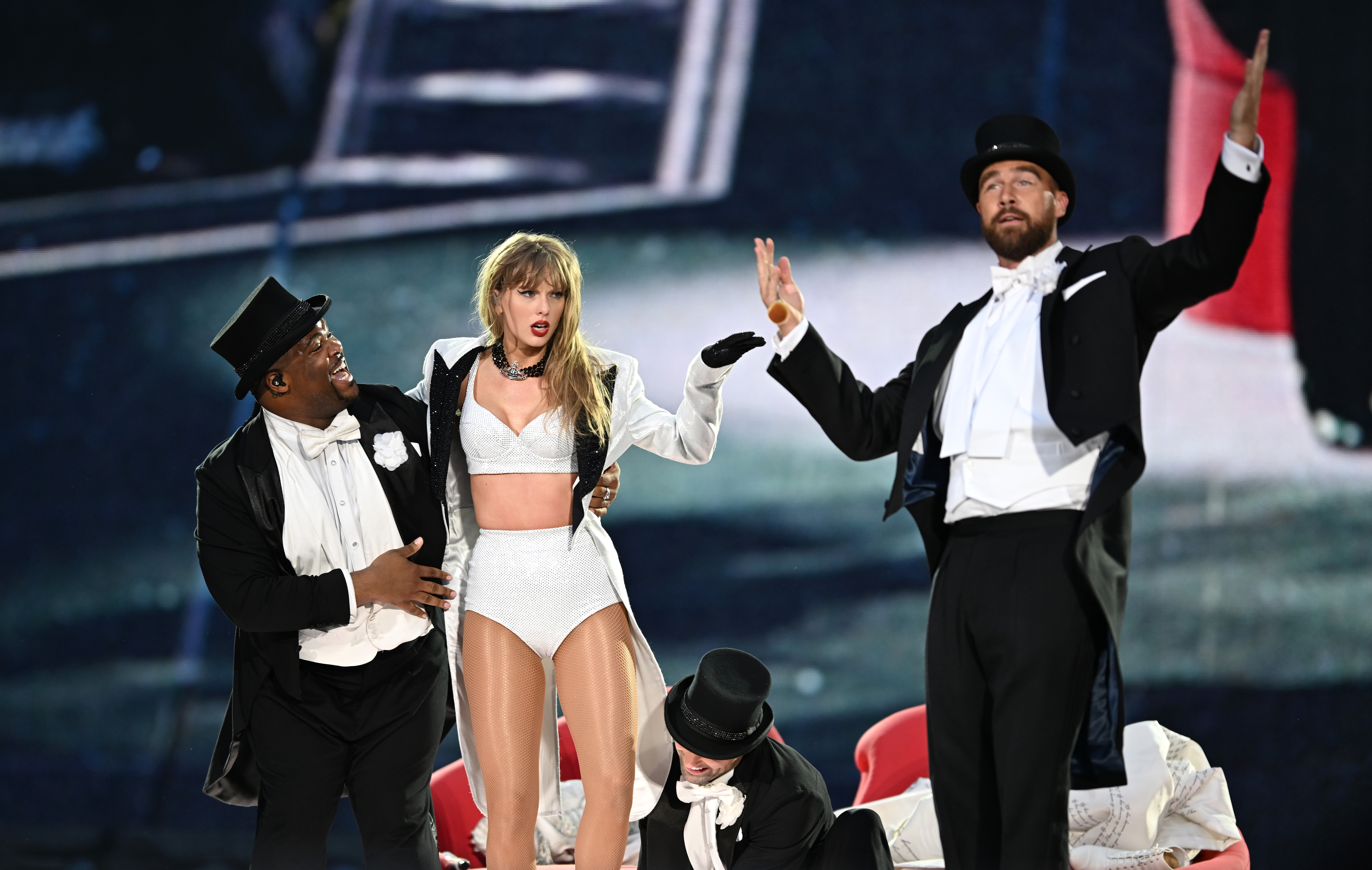 Taylor Swift performs in a white sequined two-piece outfit as Travis Kelce, dressed in a black tuxedo with a top hat, joins her on stage during "Taylor Swift | The Eras Tour" at Wembley Stadium on June 23, 2024, in London, England.