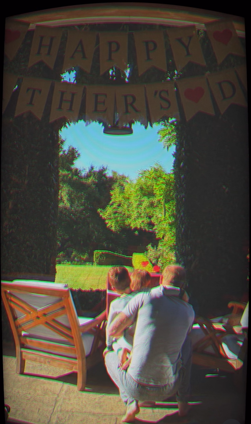 Prince Harry kneels with Prince Archie and Princess Lilibet beneath a festive Father's Day banner overlooking a sunlit garden. | Source: Instagram/meghan