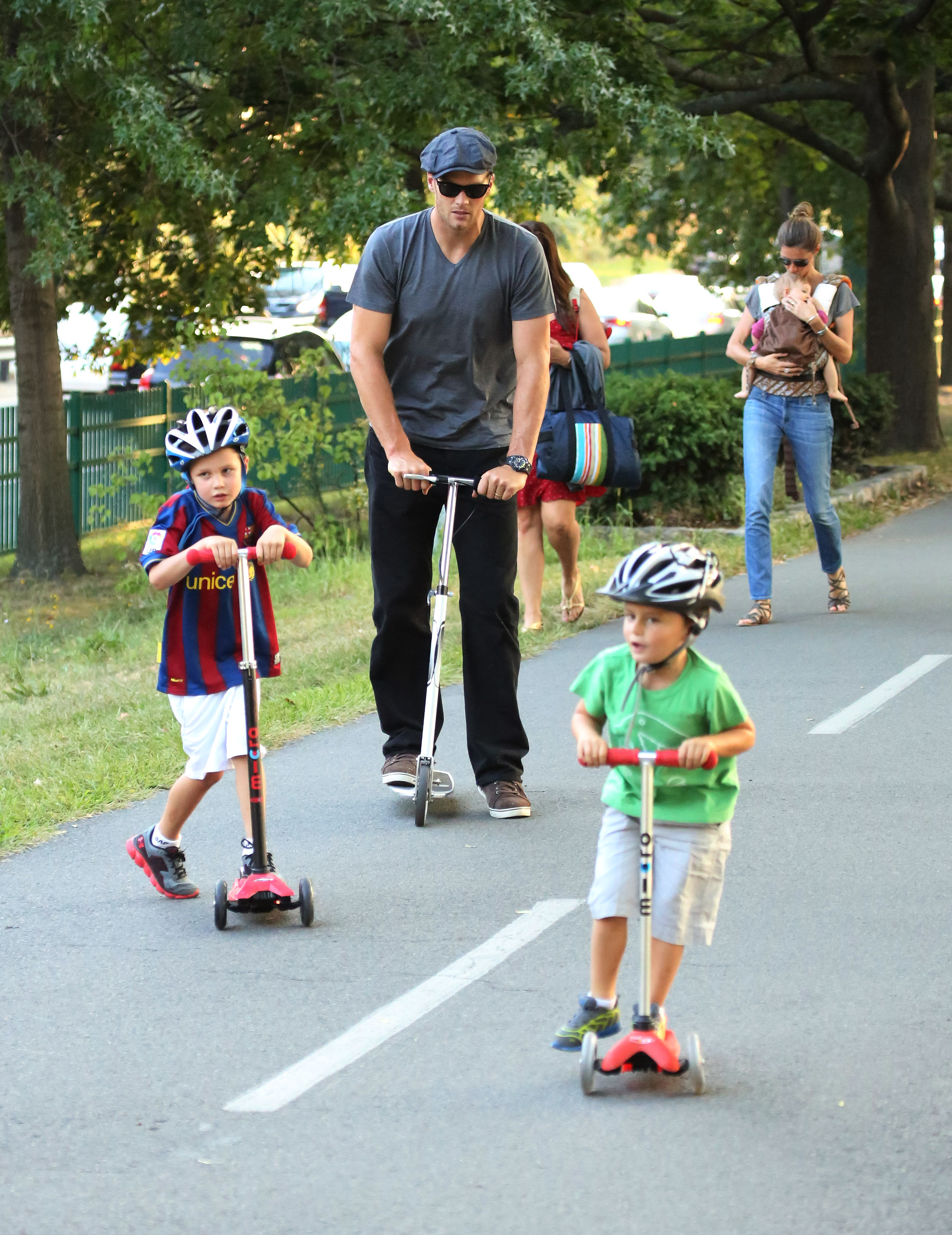 Tom Brady is seen in Boston with Jack Moynahan and Benjamin Brady on scooters, while Gisele Bündchen follows behind carrying baby Vivian Brady, making it a true family day out.
