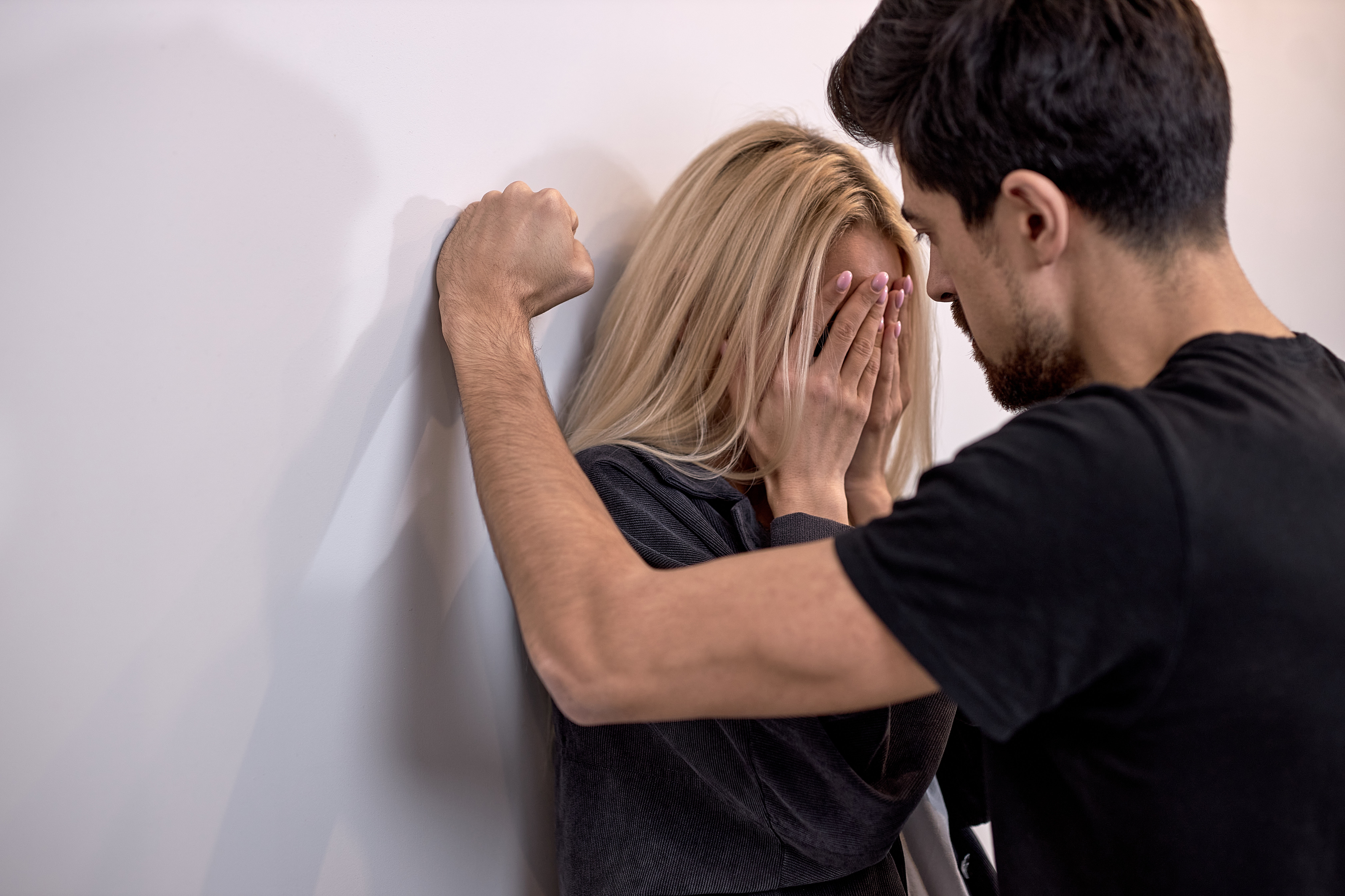 A woman standing against a wall hides her face while a man stands in front of her with his arm on the wall | Source: Shutterstock