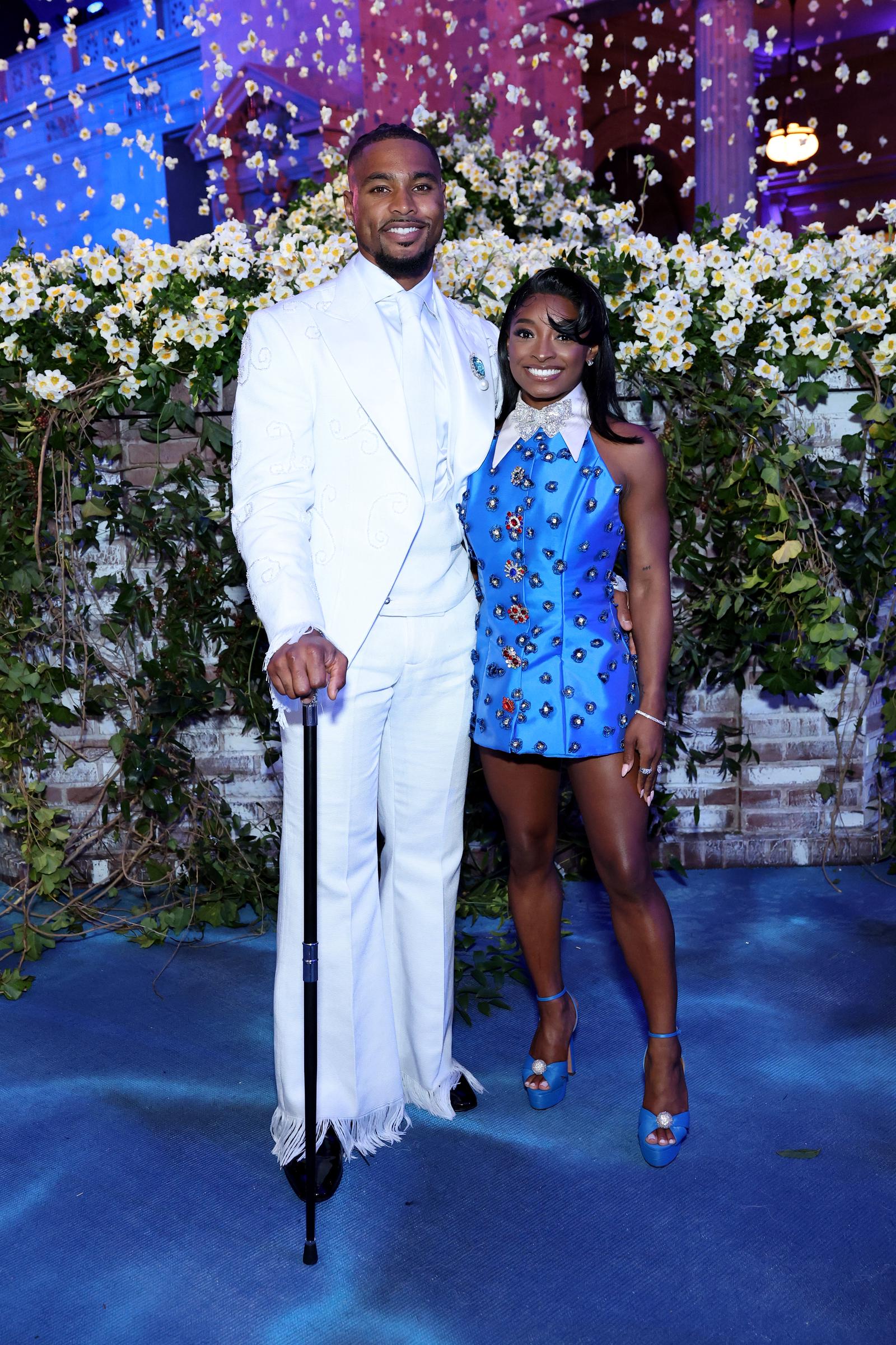 Simone Biles wears a royal blue mini dress adorned with gem-like embellishments and a dramatic oversized train, paired with matching platform heels. Beside her, Jonathan Owens stands in a textured white suit with flared hems, a white fur sash, a crystal brooch, and a black cane.