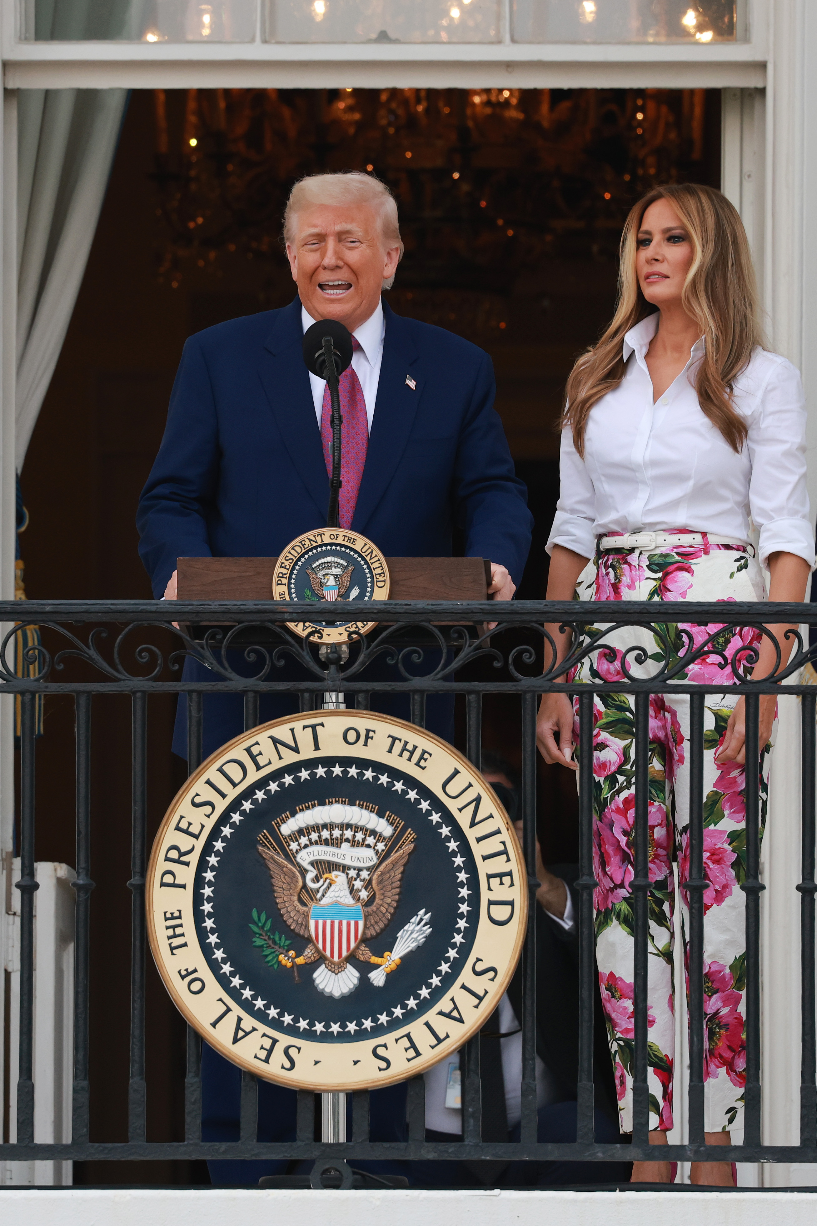 Donald Trump delivers remarks from the White House balcony as Melania stands beside him. With the presidential seal below and formal decor behind, the image captures a ceremonial moment during the 2025 Congressional Picnic.