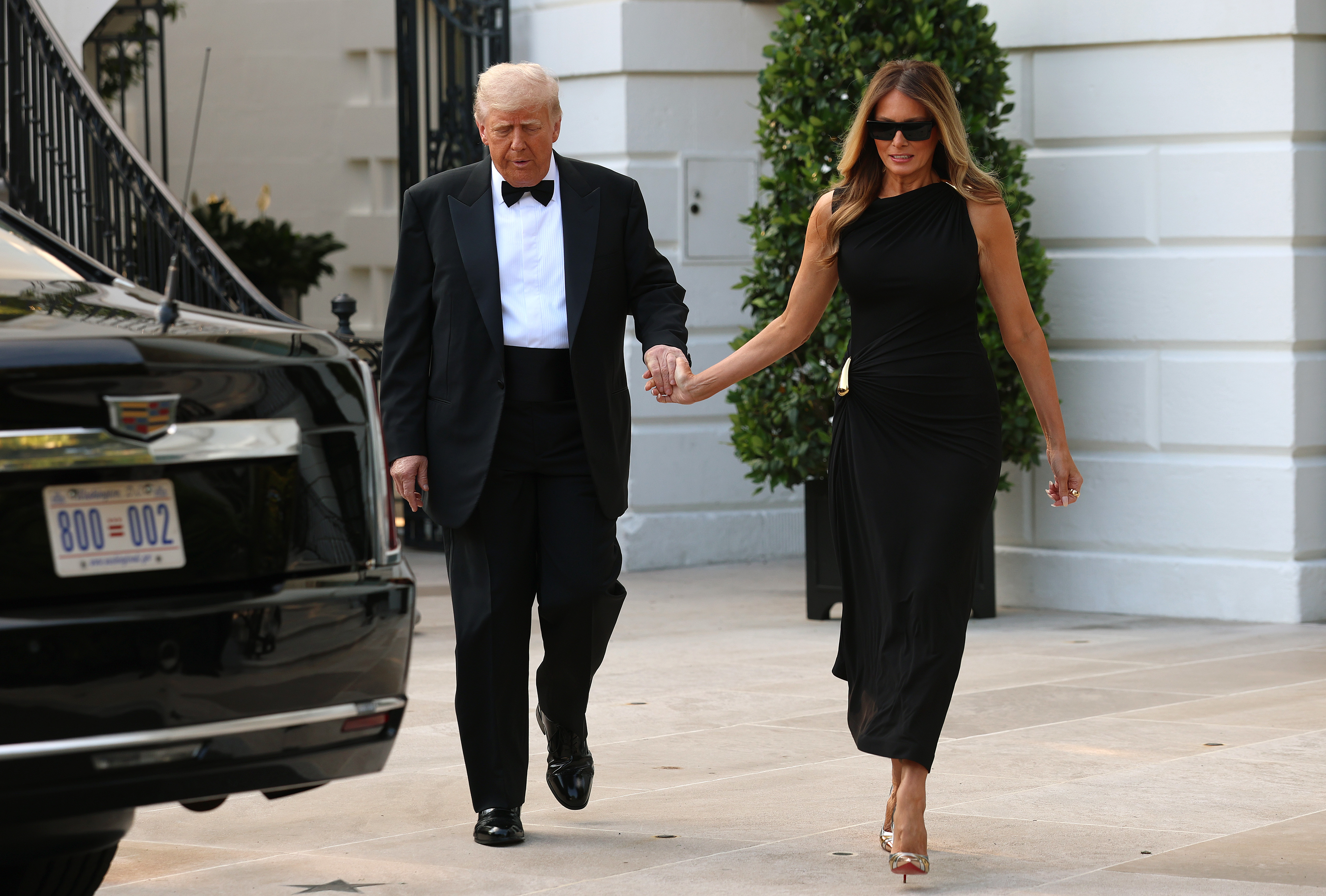 Donald and Melania walk hand in hand in front of the White House, approaching a waiting vehicle. Melania leads slightly ahead, her black dress flowing as she steps forward in metallic heels. Her sunglasses and confident stride give her a striking, modern elegance.