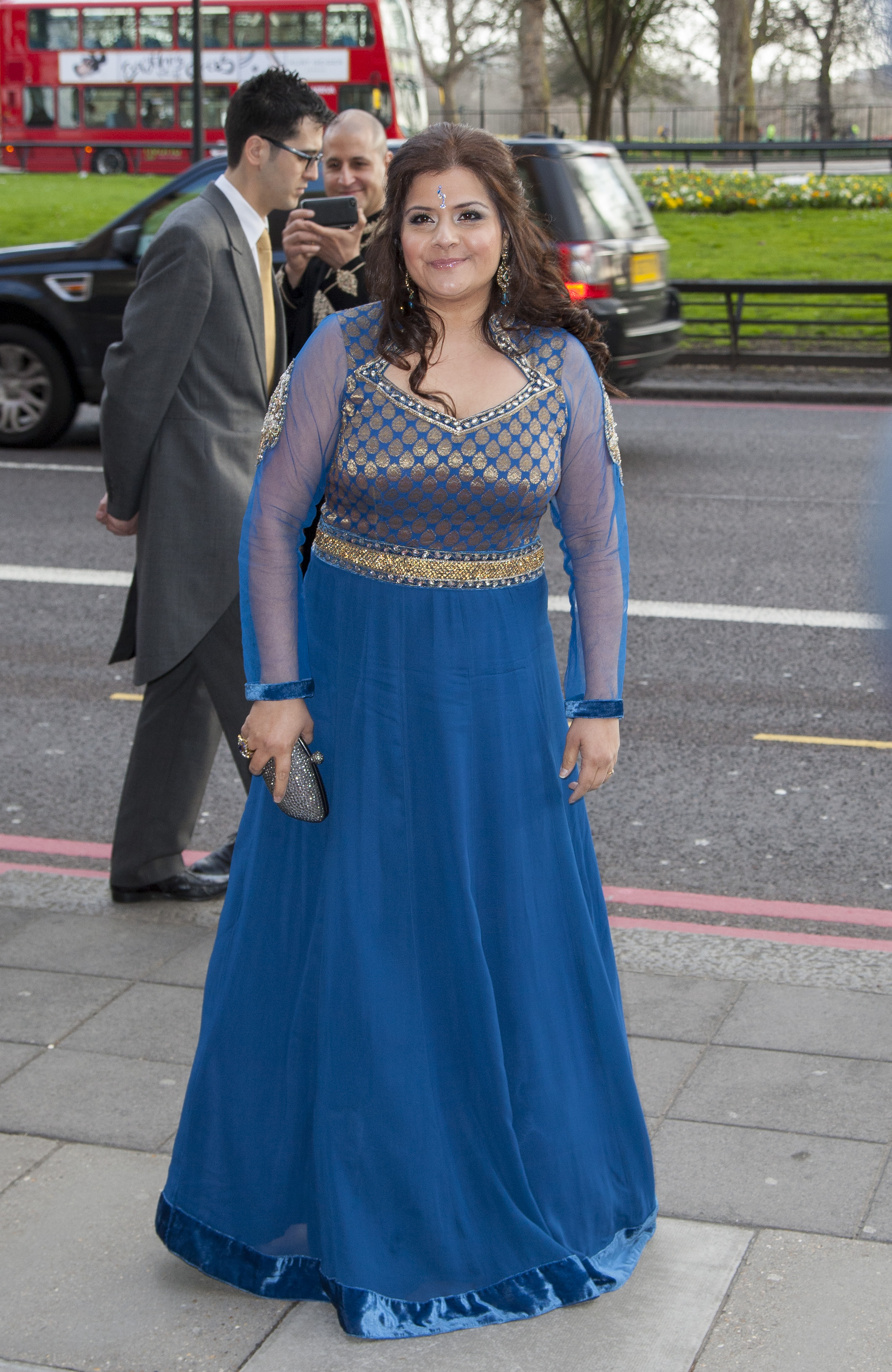 Wadia arrives at The Asian Awards at Grosvenor House in April 2013 wearing a sweeping cobalt blue anarkali gown with sheer mesh sleeves and an intricately embroidered gold and blue bodice. A delicate bindi sits at her hairline, and she carries a small silver crystal clutch, with a red London double-decker bus visible on the street behind her.