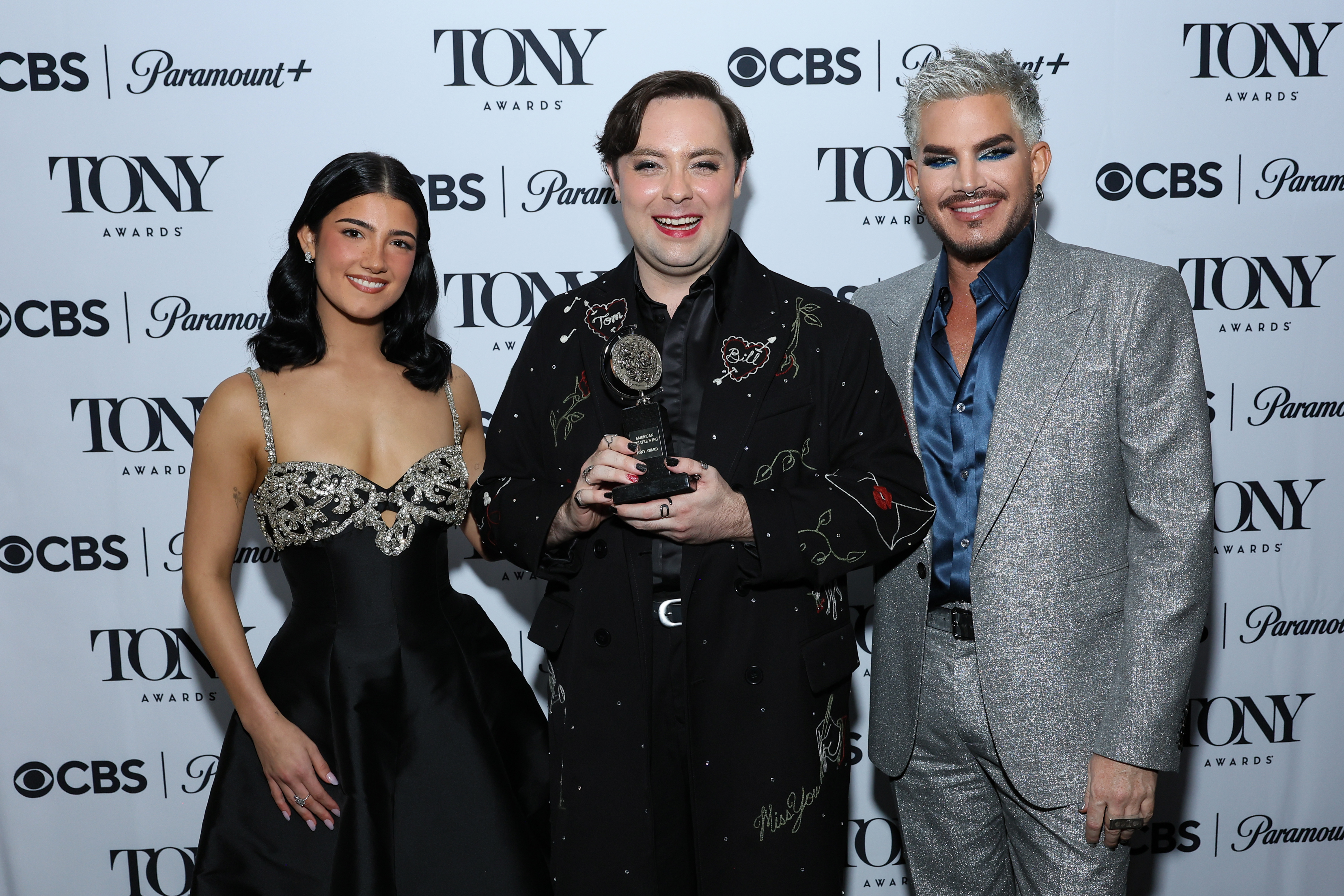 Backstage, Adam Lambert poses with Charli D’Amelio and award-winner Jak Malone. Their smiles, the metallic sheen of Lambert's suit, and Malone's trophy at the center create a celebratory snapshot after a standout award presentation.