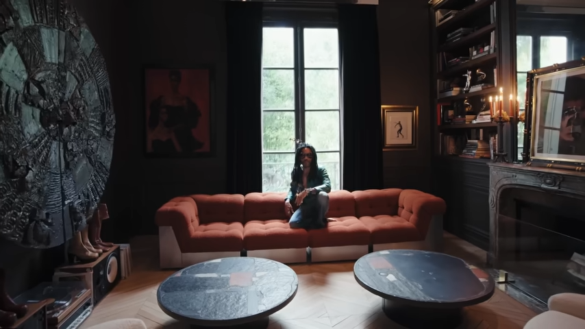 Lenny Kravitz sits on his comfortable rust-colored modular sofa in his library, framed by a massive circular metal wall sculpture and the room's high arched windows. | Source: YouTube/@Archdigest