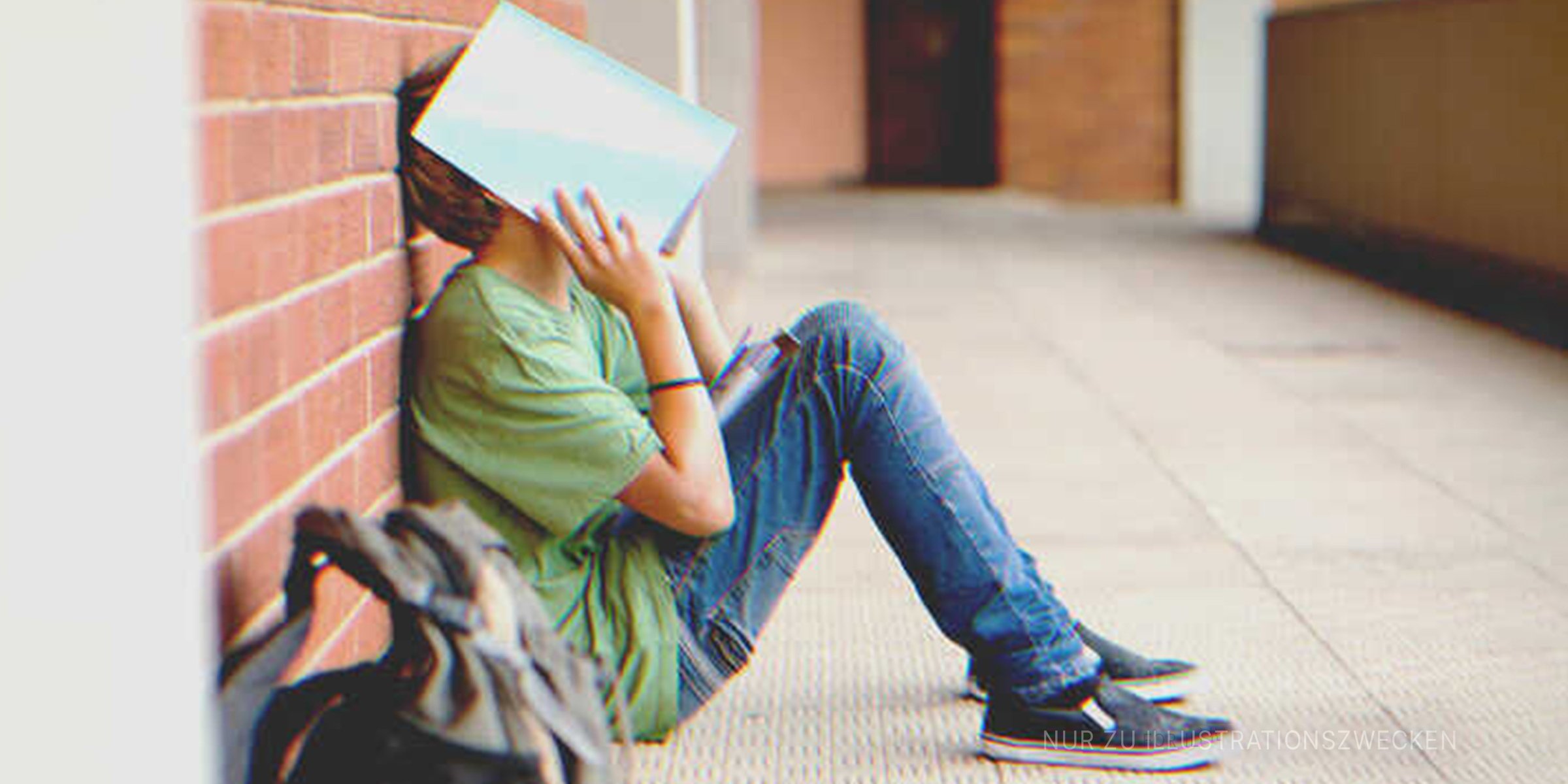 Schuljunge sitzt außerhalb des Klassenzimmers mit einem Buch, das sein Gesicht bedeckt | Quelle: Getty Images