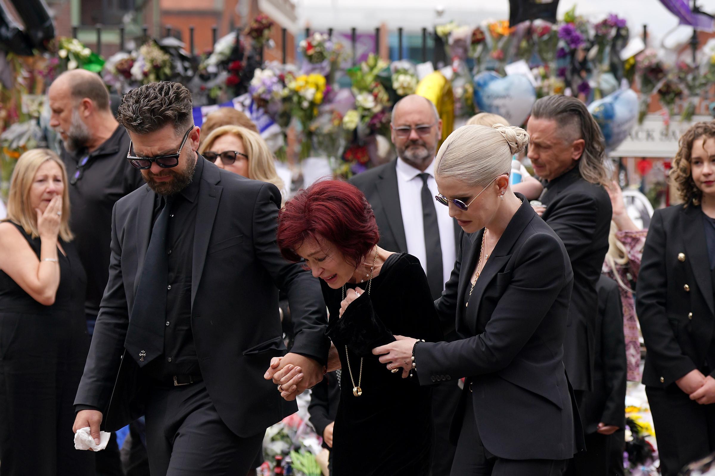 Jack, Sharon, and Kelly Osbourne move slowly together as they hold hands while approaching the messages and floral tributes left at the Black Sabbath Bench and Bridge on Broad Street in Birmingham on July 30, 2025. Surrounded by mourners and memorials, the family appears visibly emotional as they support one another during the procession.