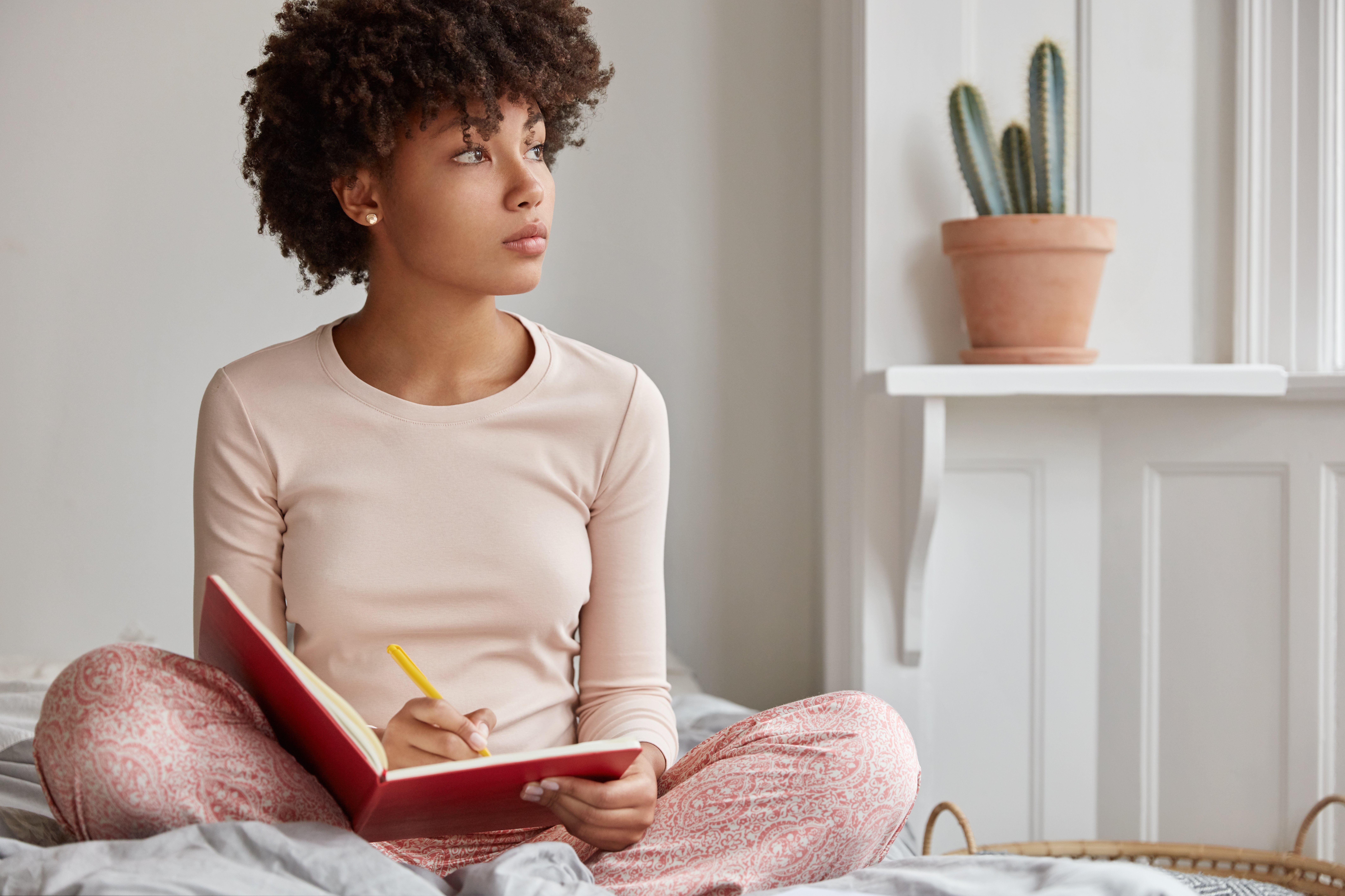 A woman sitting cross-legged on her bed, looking out the window as she journals | Source: Shutterstock