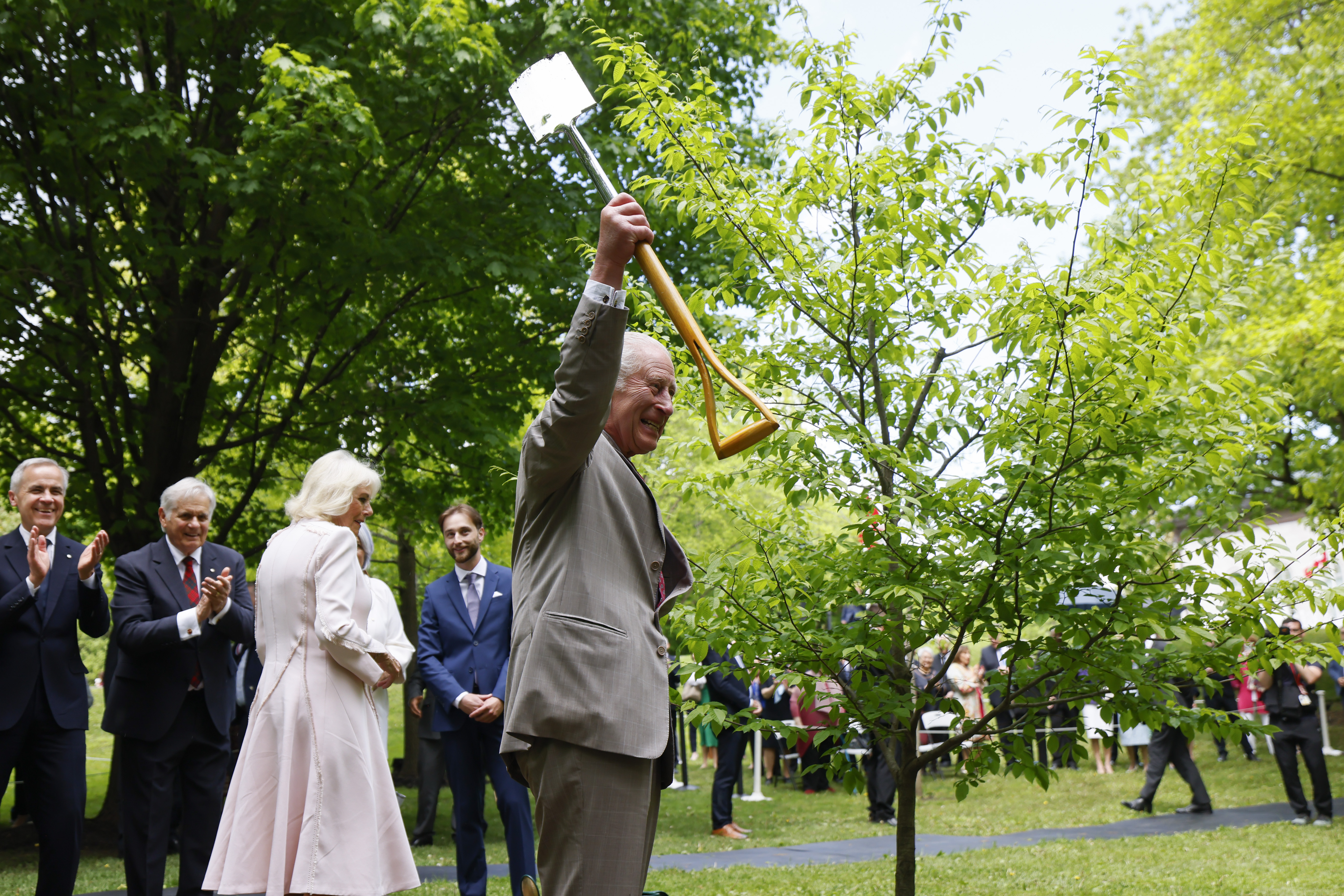 King Charles III holds a shovel as he joins Queen Camilla in a ceremonial tree planting at Rideau Hall. Surrounding them, officials — including Prime Minister Mark Carney — applaud warmly after the tree is planted, marking a moment of celebration and symbolism.