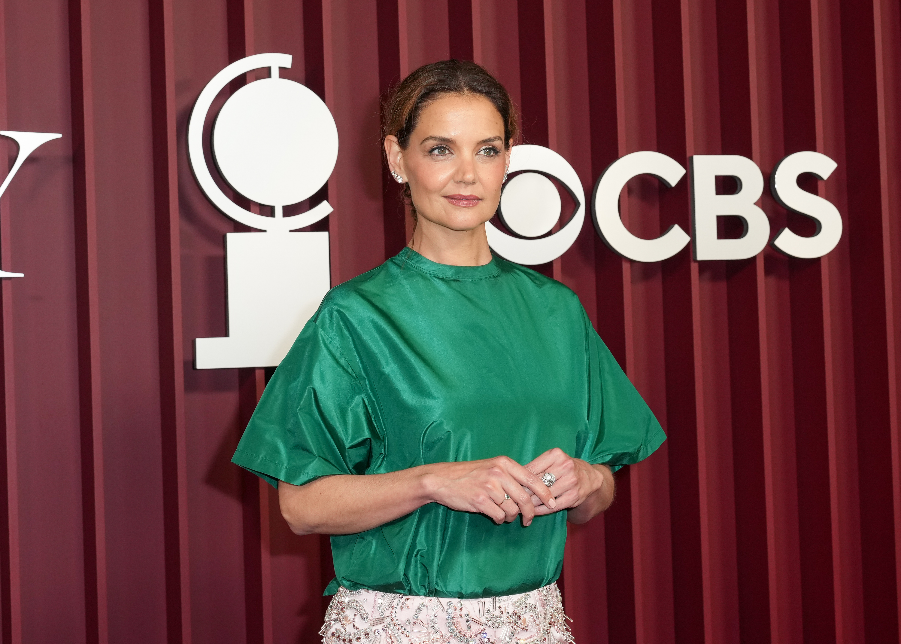 Wearing a bold color combo, Katie Holmes appears poised in green satin and soft-toned makeup, standing confidently in front of the CBS and Tony logos. Her sleek low bun and subtle jewelry complete the understated red carpet look.