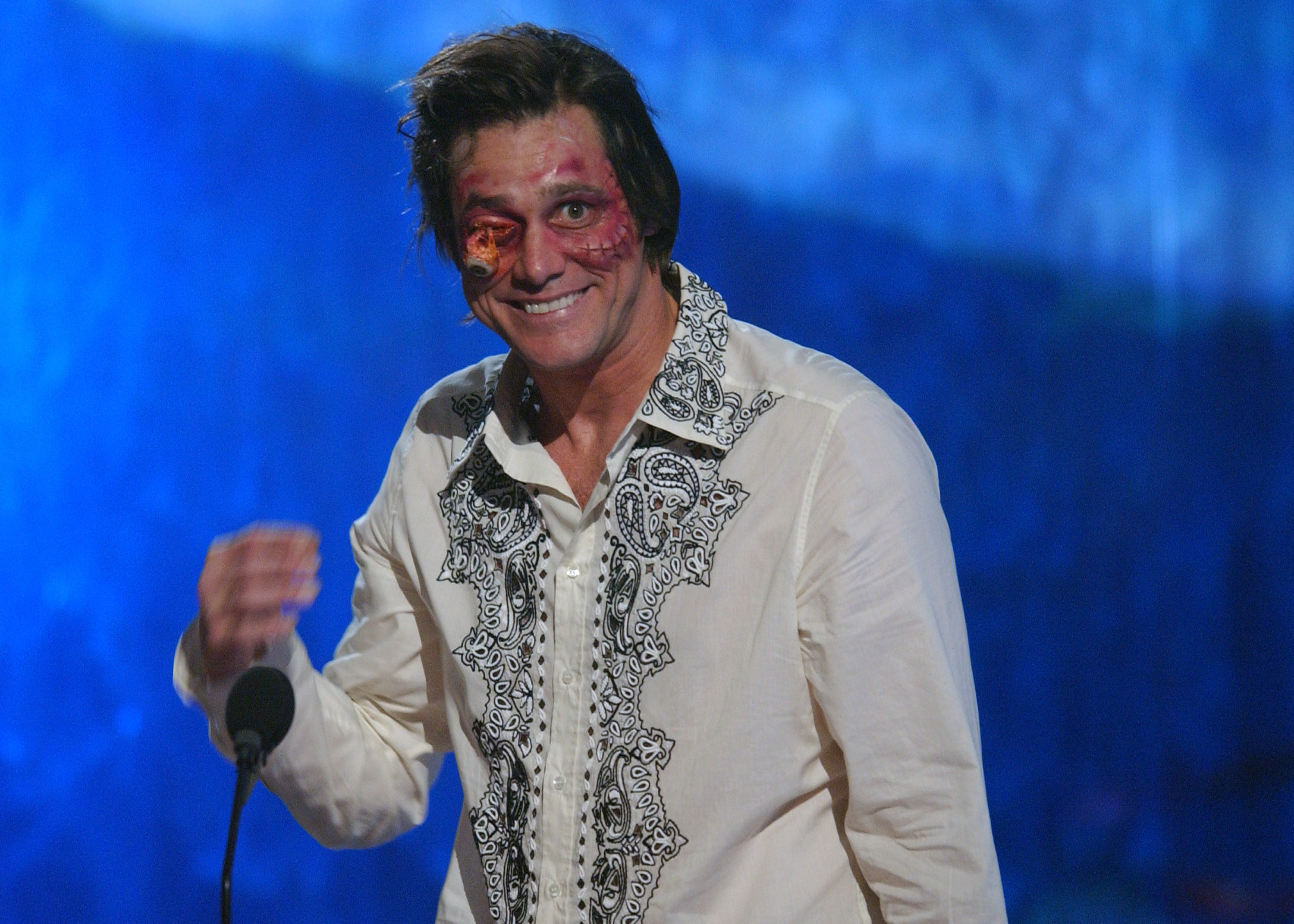 At the Teen Choice Awards stage, Jim Carrey wears a white embroidered shirt and sports dramatic special-effects makeup on his face. Despite the exaggerated, humorous “injuries,” he smiles broadly, embodying his trademark comedic flair. The vibrant blue backdrop adds to the lively and playful atmosphere of the moment.