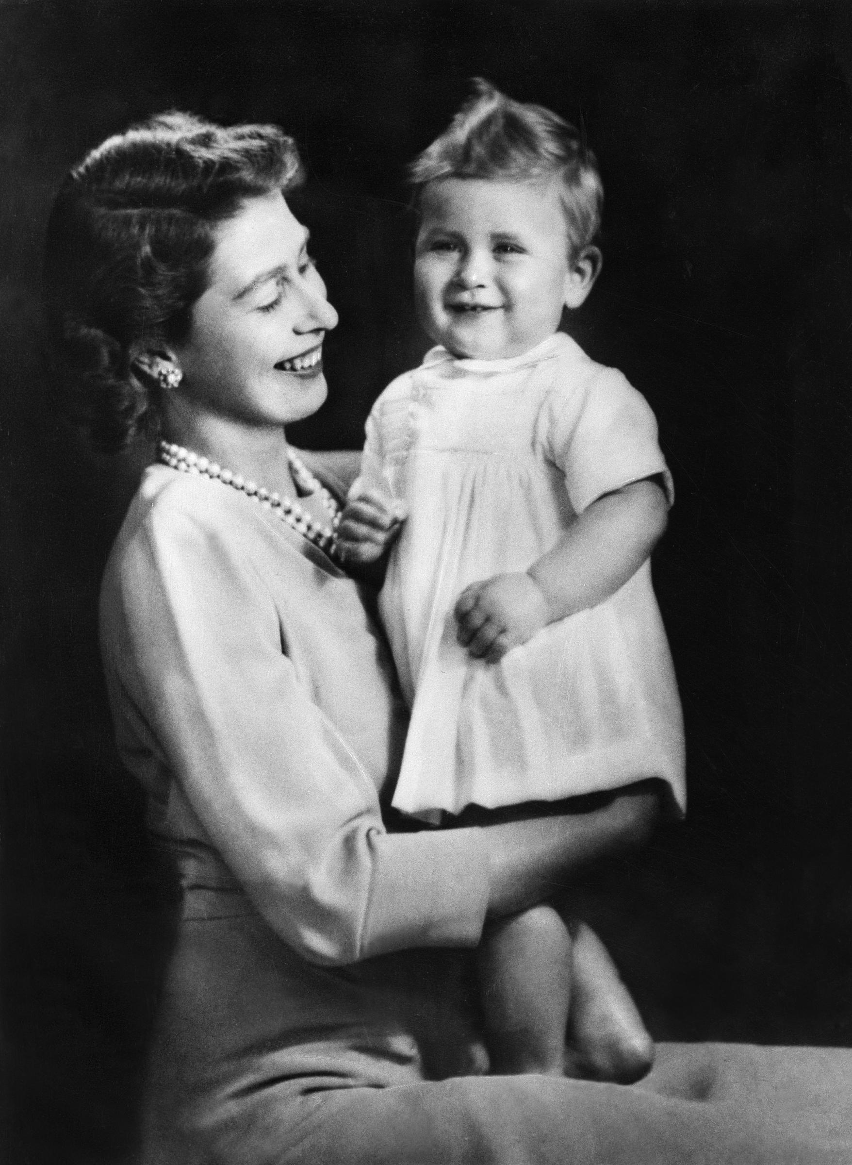 In a striking half-portrait, the future Queen Elizabeth II is seen smiling warmly as she holds a young Prince Charles in her arms, the candid moment capturing a sense of maternal joy and intimacy as the toddler beams with delight, offering a timeless glimpse into their early bond before her coronation in 1953.