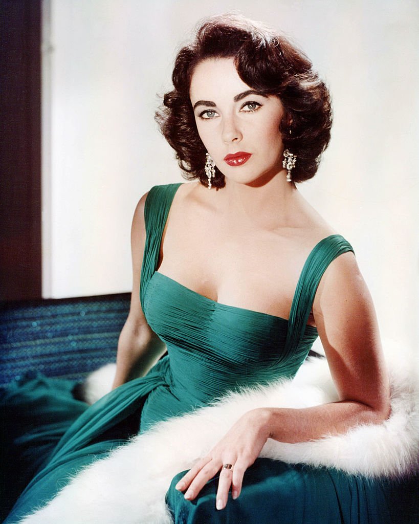 Elizabeth Taylor wearing a green sleeveless low-cut dress, with a white fur wrap on the arm of the armchair in which she sits, captured in 1950 | Photo: Getty Images