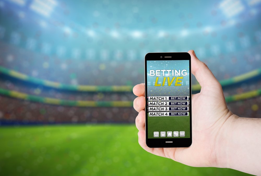 Mano sostiene un teléfono con apuestas en línea en una pantalla en el fondo de un estadio. I Foto: Shutterstock Mano sostiene un teléfono con apuestas en línea en una pantalla en el fondo de un estadio. I Foto: Shutterstock