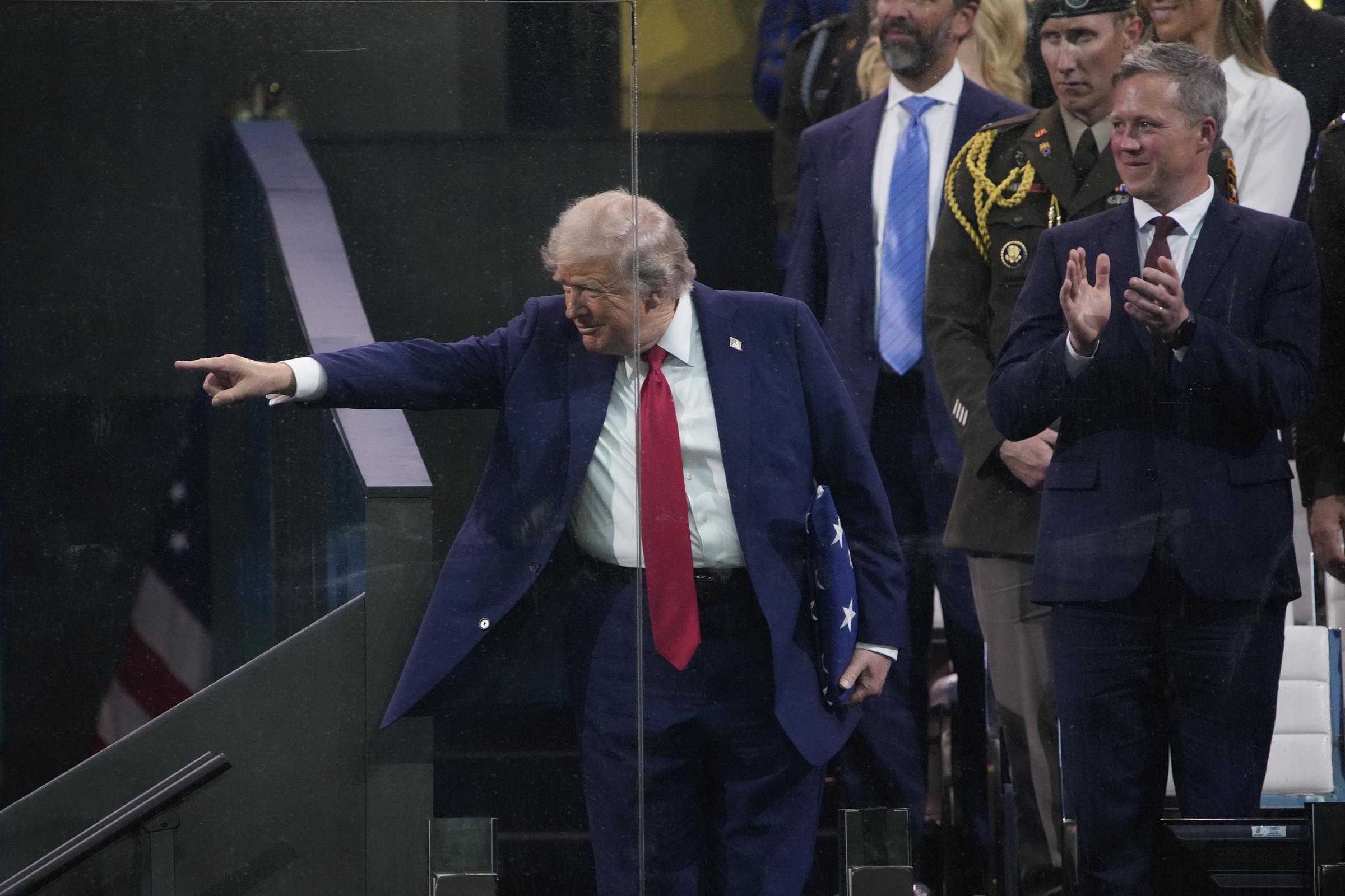 Donald Trump points toward the audience after receiving a folded flag, while guests behind him applaud. His gesture adds a spontaneous moment during an otherwise structured military celebration.