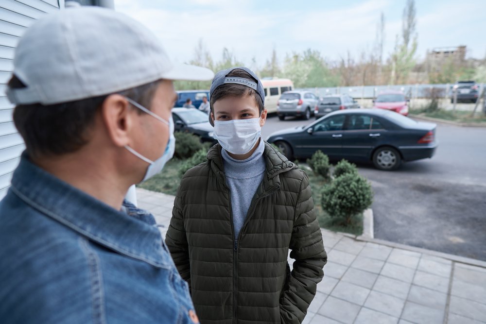 A man talks to a boy near a wall and closed-door while both wear medical masks on their faces | Photo: Shutterstock/s_oleg