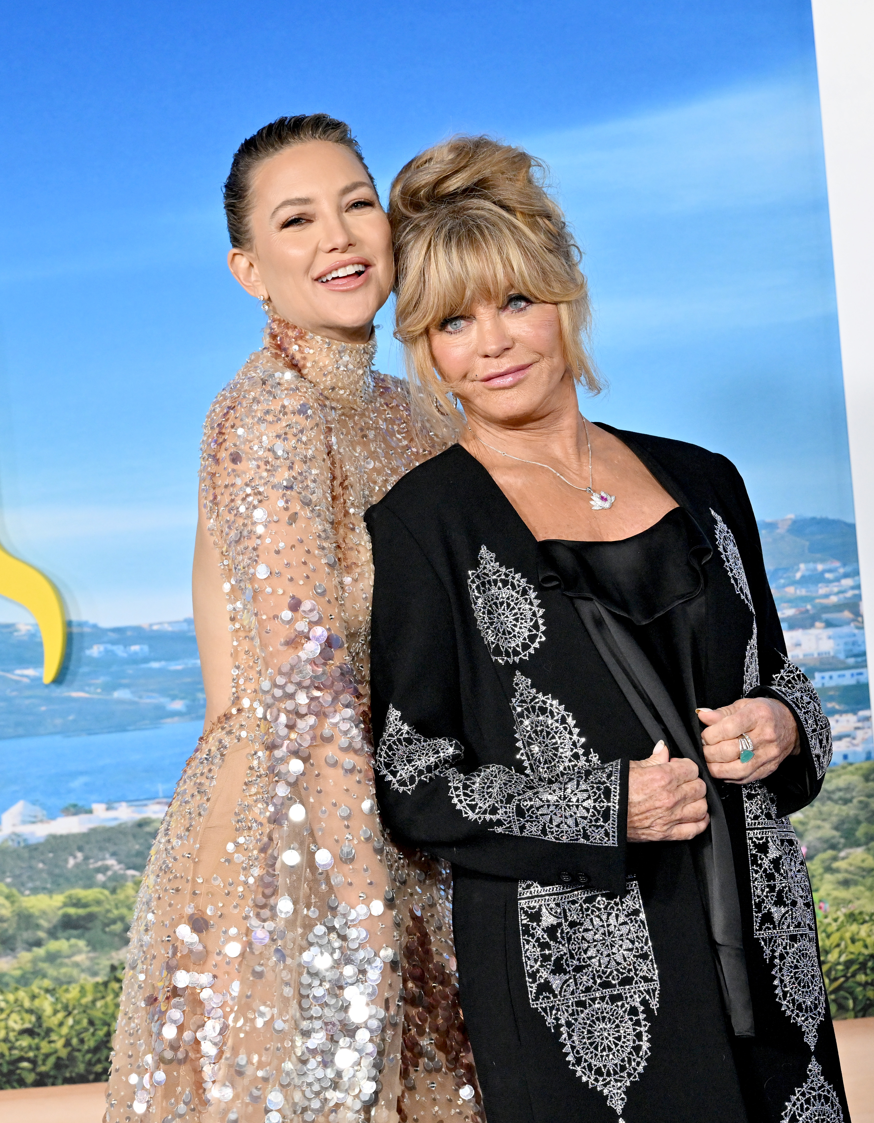 At the "Glass Onion: Knives Out" premiere, Kate Hudson and Goldie Hawn pose in contrasting styles. Hudson glows in a nude, beaded gown with slicked-back hair, while Hawn complements her in a long black coat adorned with silver embroidery.
