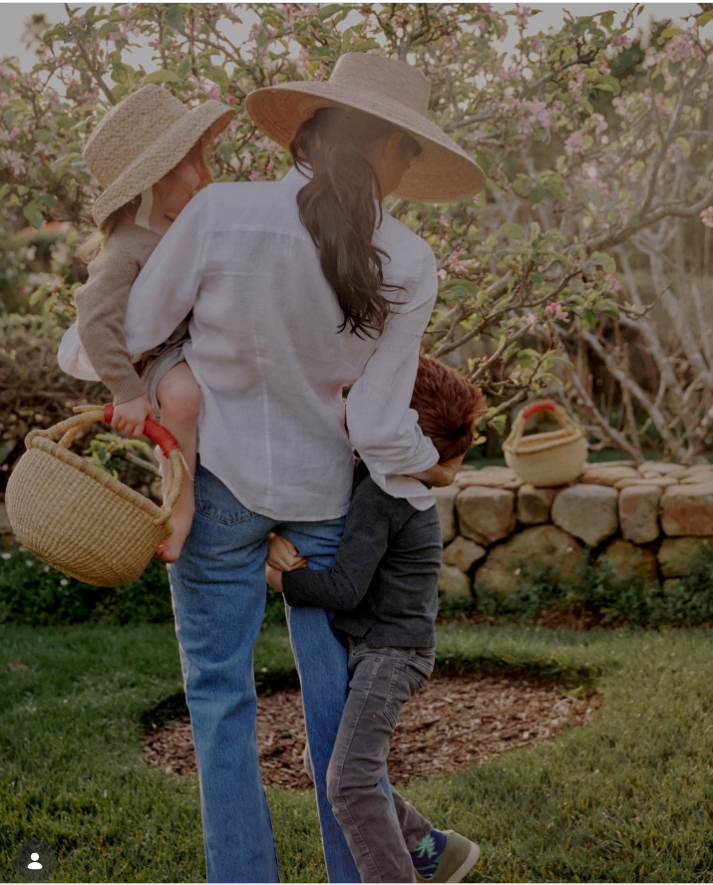 Meghan Markle walks through a blooming orchard carrying one child while another clings to her side, capturing a candid moment of family life outdoors. | Source: aseverofficial and meghan